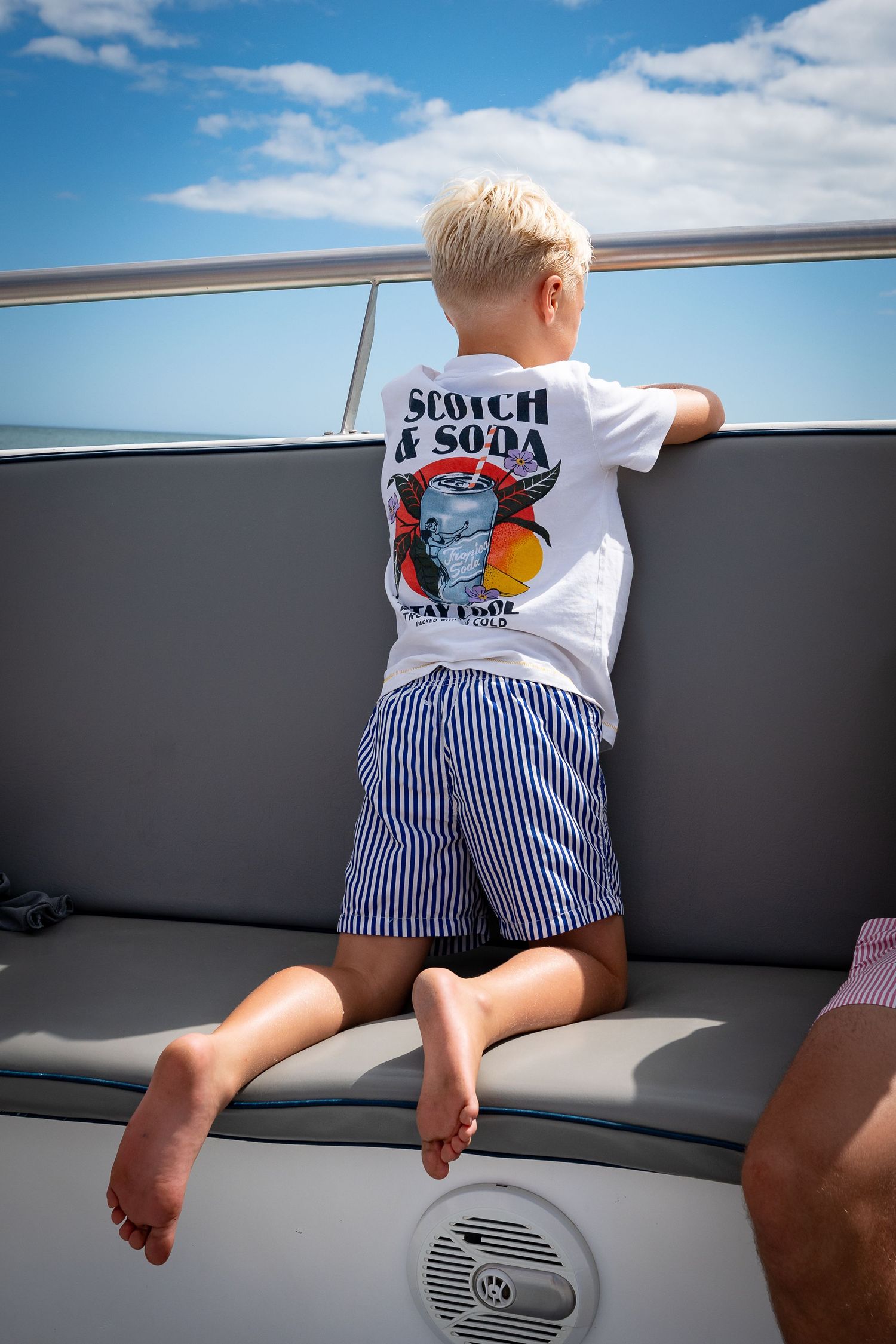 Young passenger in striped shorts and graphic t-shirt kneeling on boat seat looking out over railing on sunny day.