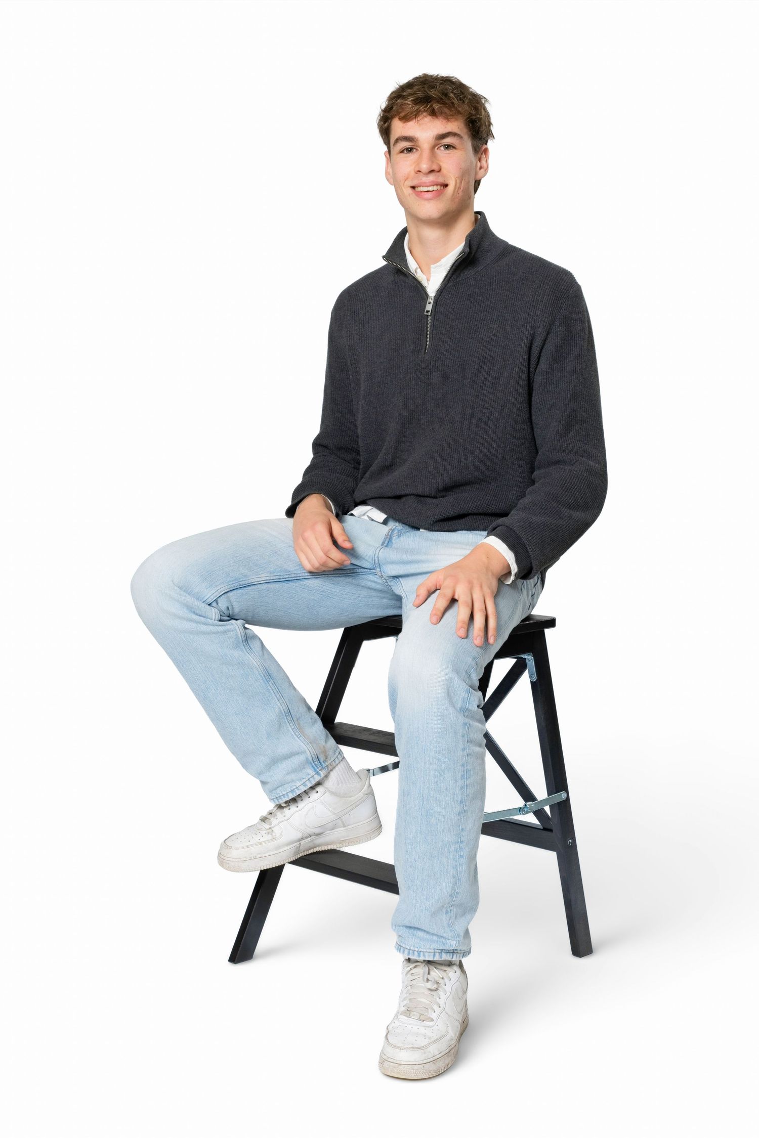 Smiling young man in a gray quarter-zip sweater, light jeans and white sneakers sitting relaxed on a black folding stool.