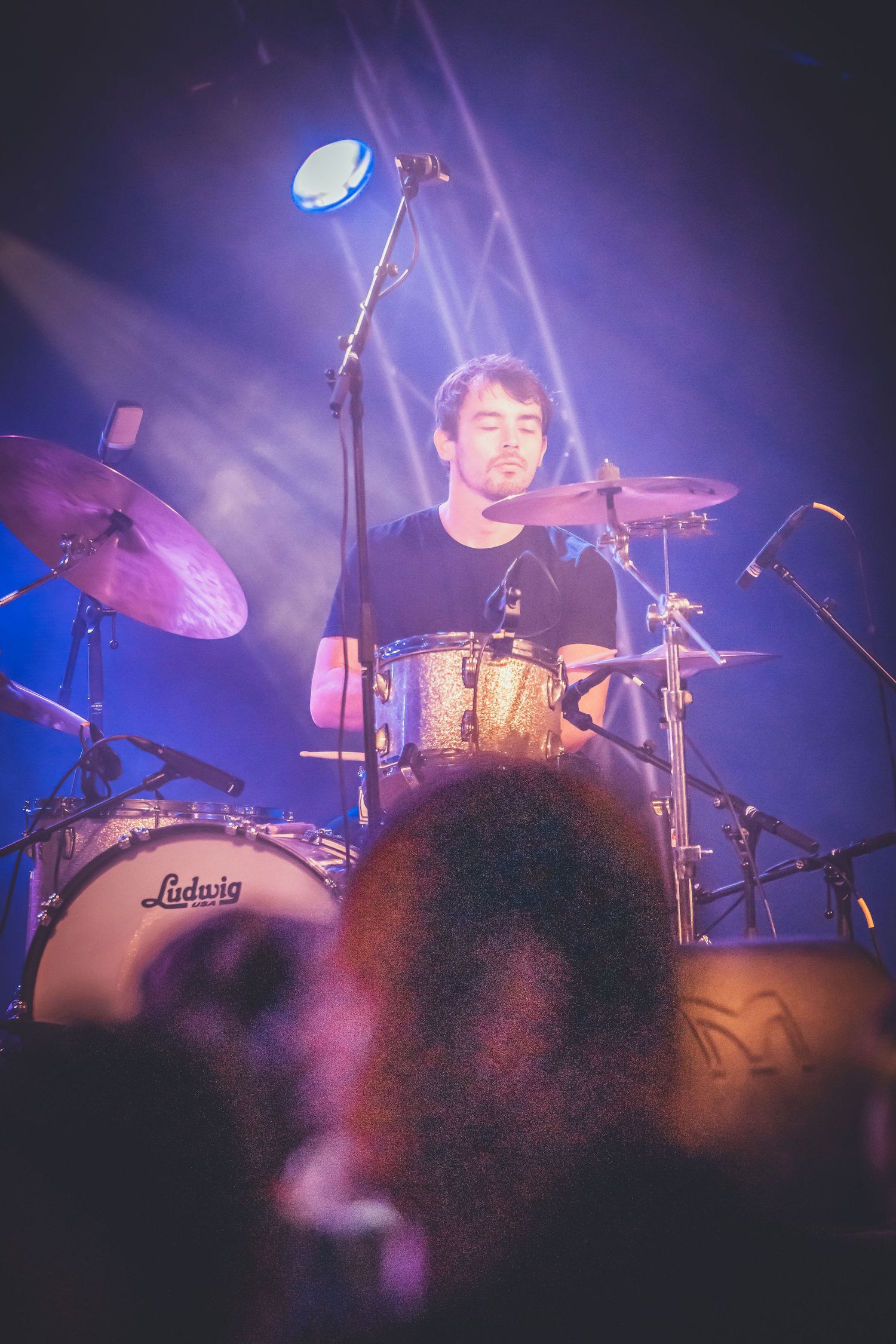 Concert drummer performing on stage with Ludwig drum kit illuminated by dramatic blue and purple stage lighting.