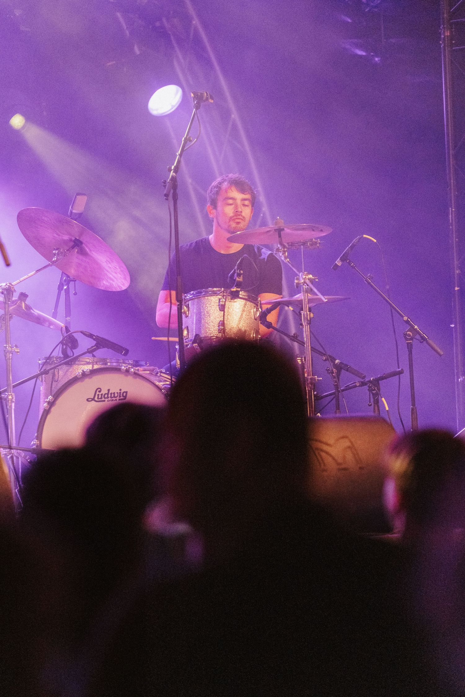 Purple stage lighting illuminates a drummer performing at a concert with silhouetted crowd in foreground.