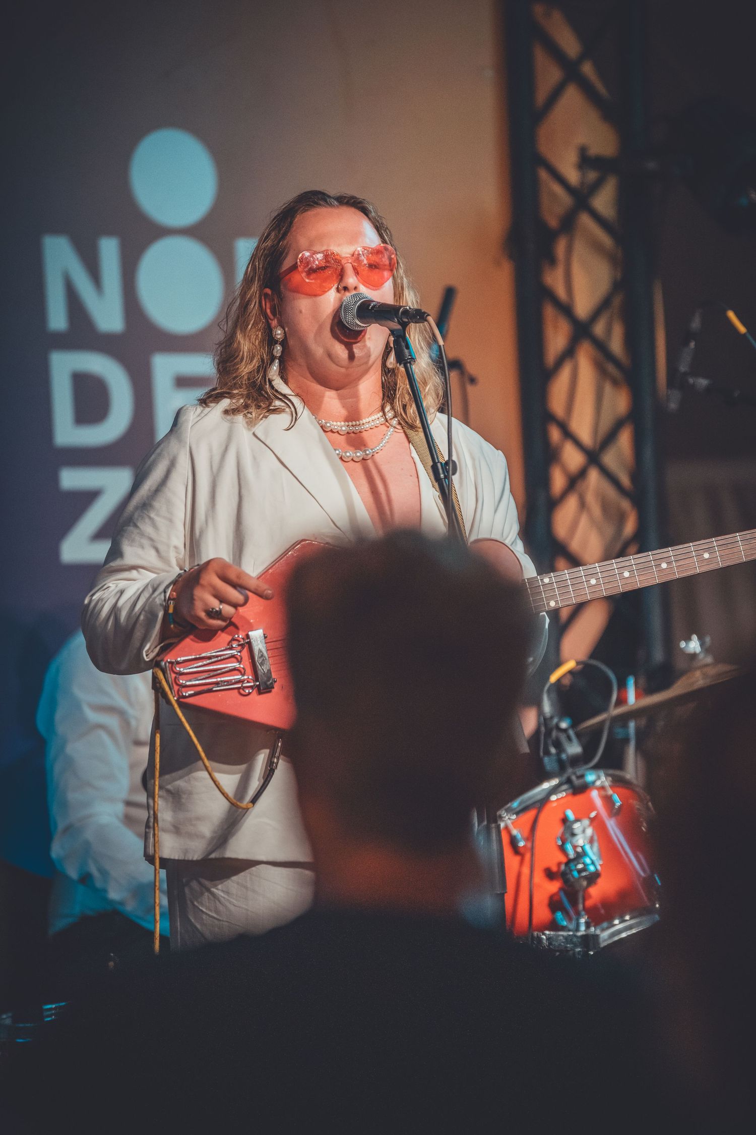 A performer in a white suit and red glasses sings into a microphone during a concert with a blue logo visible in background.