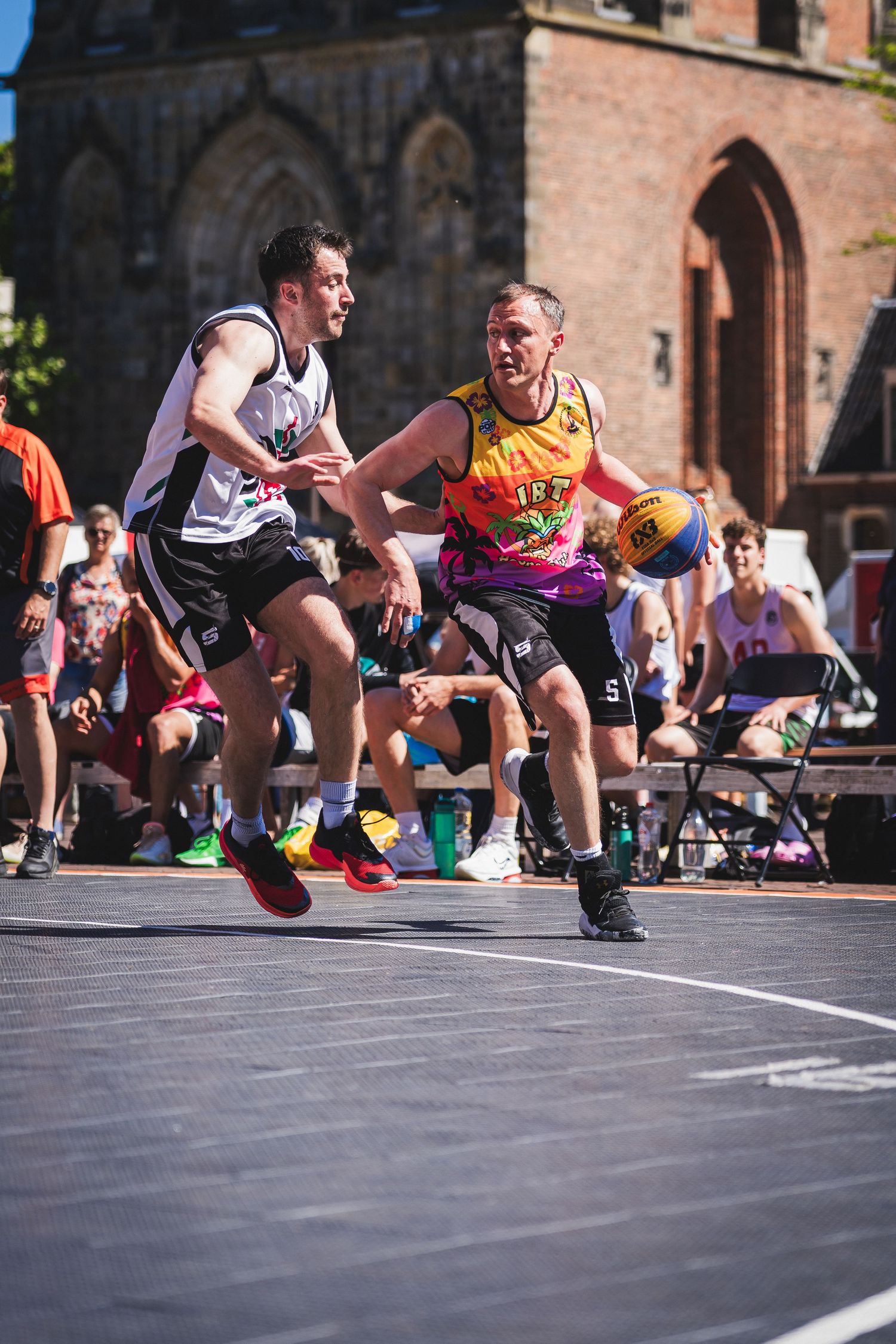 Two male players compete in an outdoor 3x3 street basketball game near a historic brick building on a sunny day.