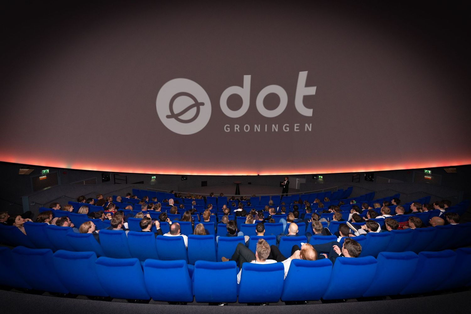 Audience wearing blue uniforms seated in a dome theater with red lighting and dot Groningen logo displayed above.