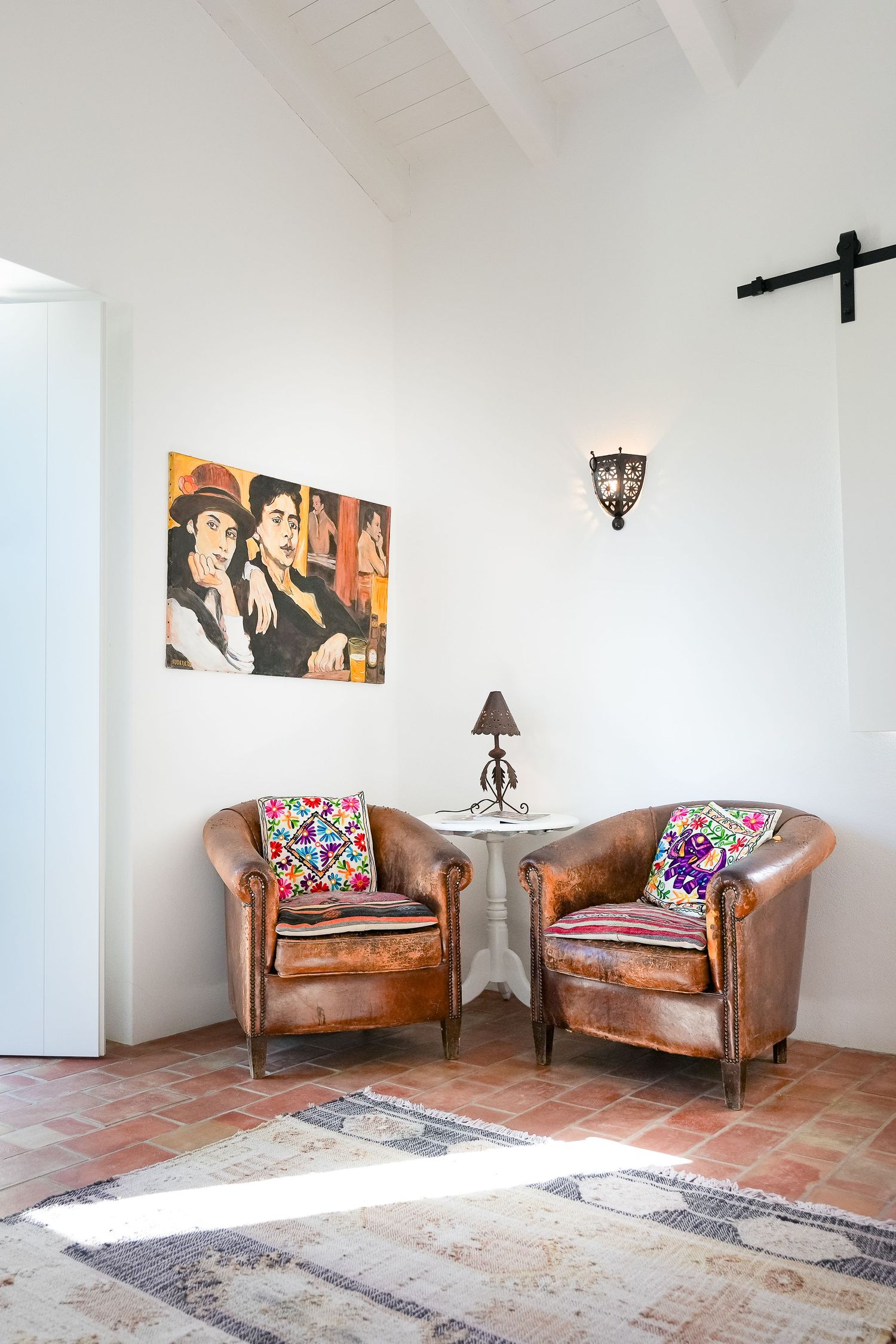 Modern living room corner with brown leather armchairs, colorful throw pillows, and vintage area rug on terracotta tile floors.