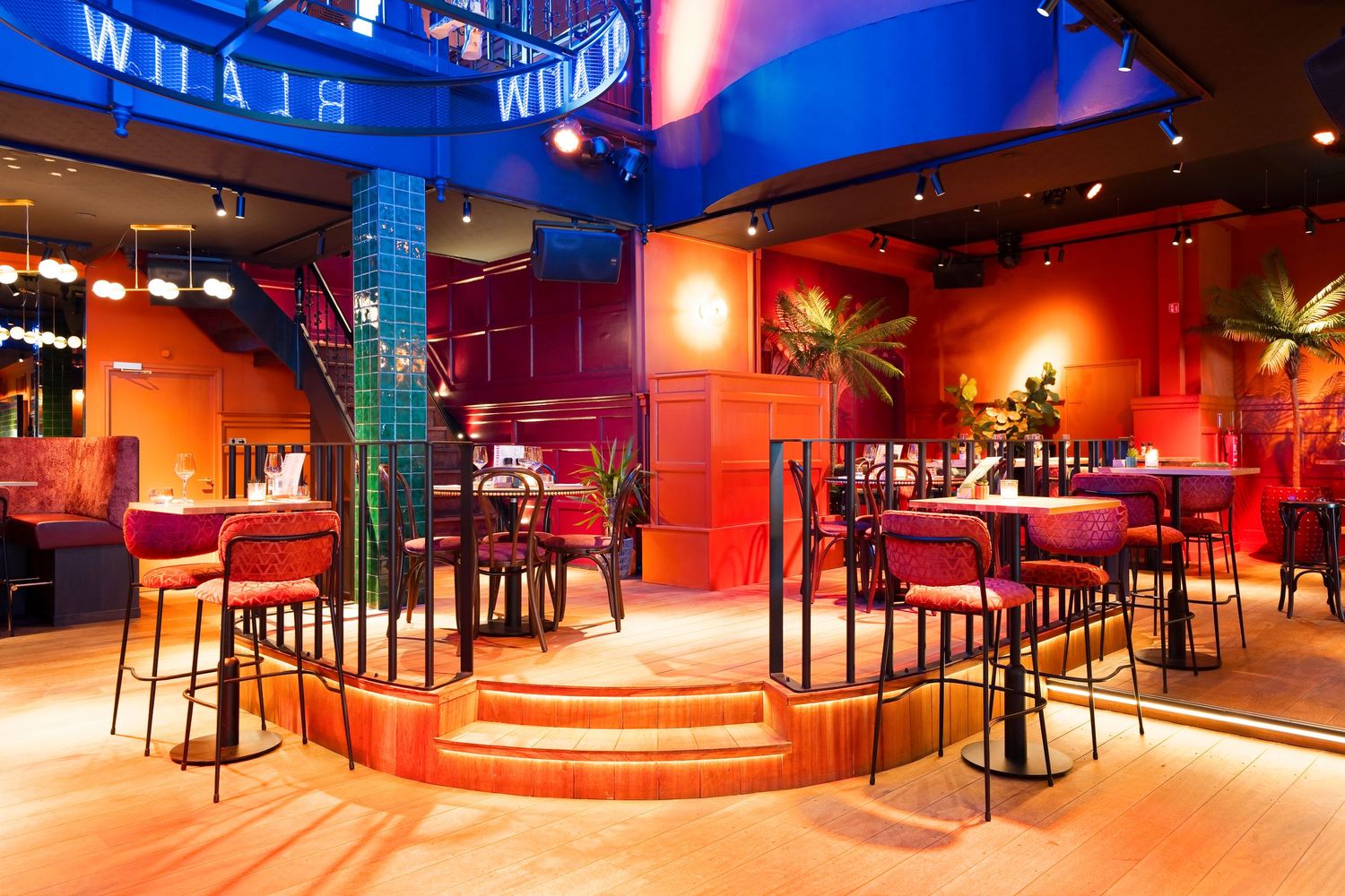 Vibrant restaurant interior with red and orange decor, high-top tables, bar seating, and tropical plants under neon signs.
