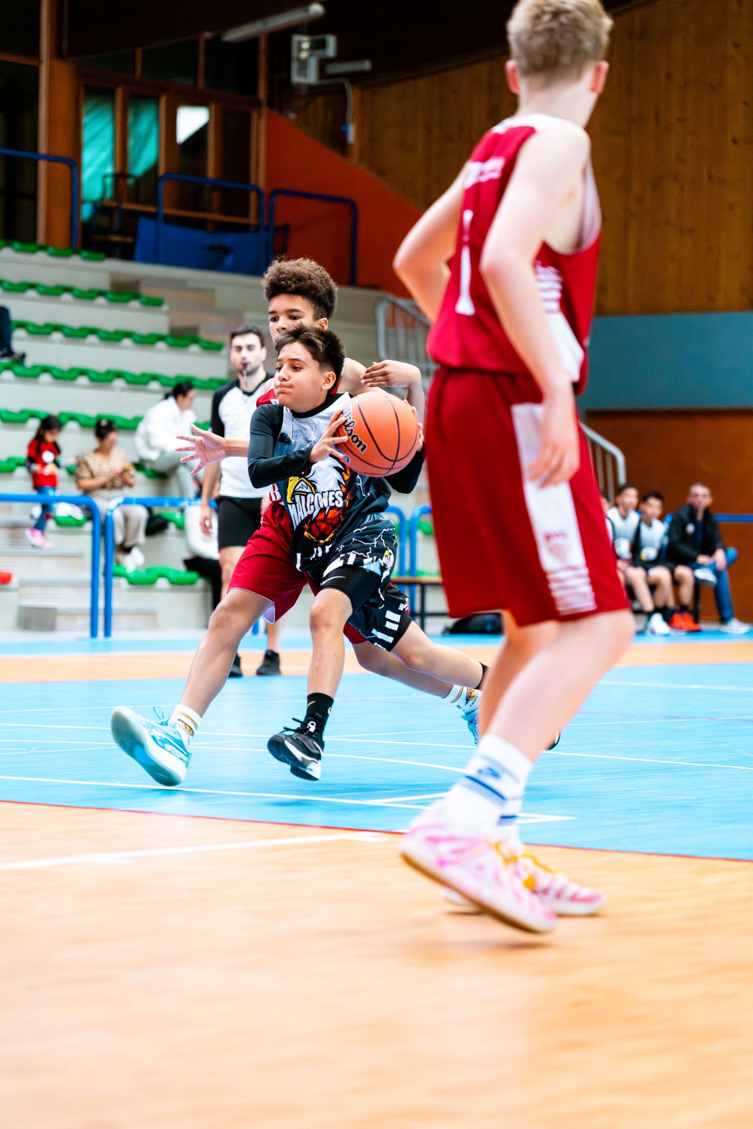 Young basketball players compete in an indoor court game, with one player driving to the basket while defenders pursue.