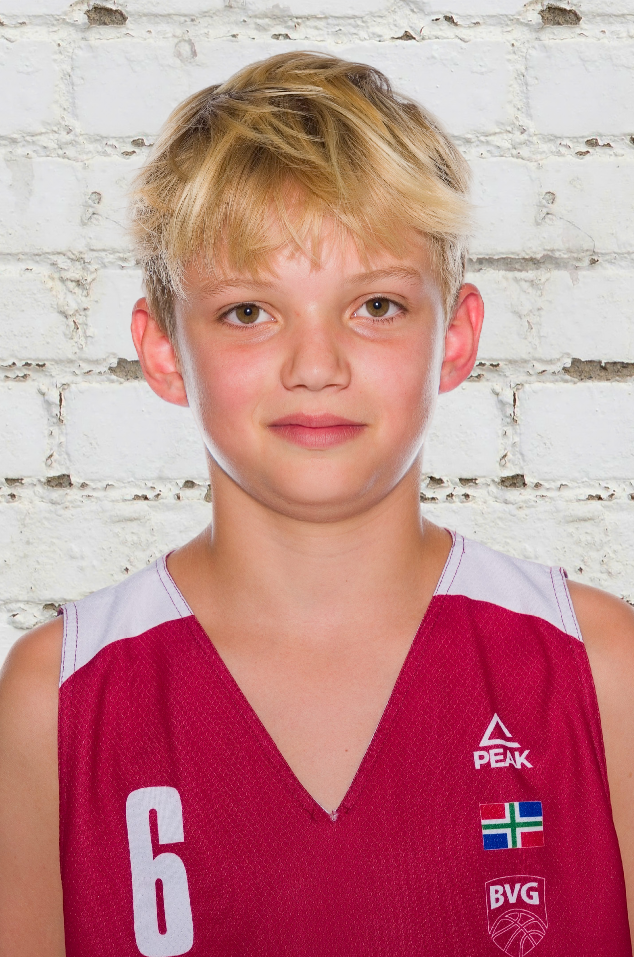 Young basketball player with blond hair wearing a red number 6 Peak jersey with BVG logo, posing against a white painted brick wall.