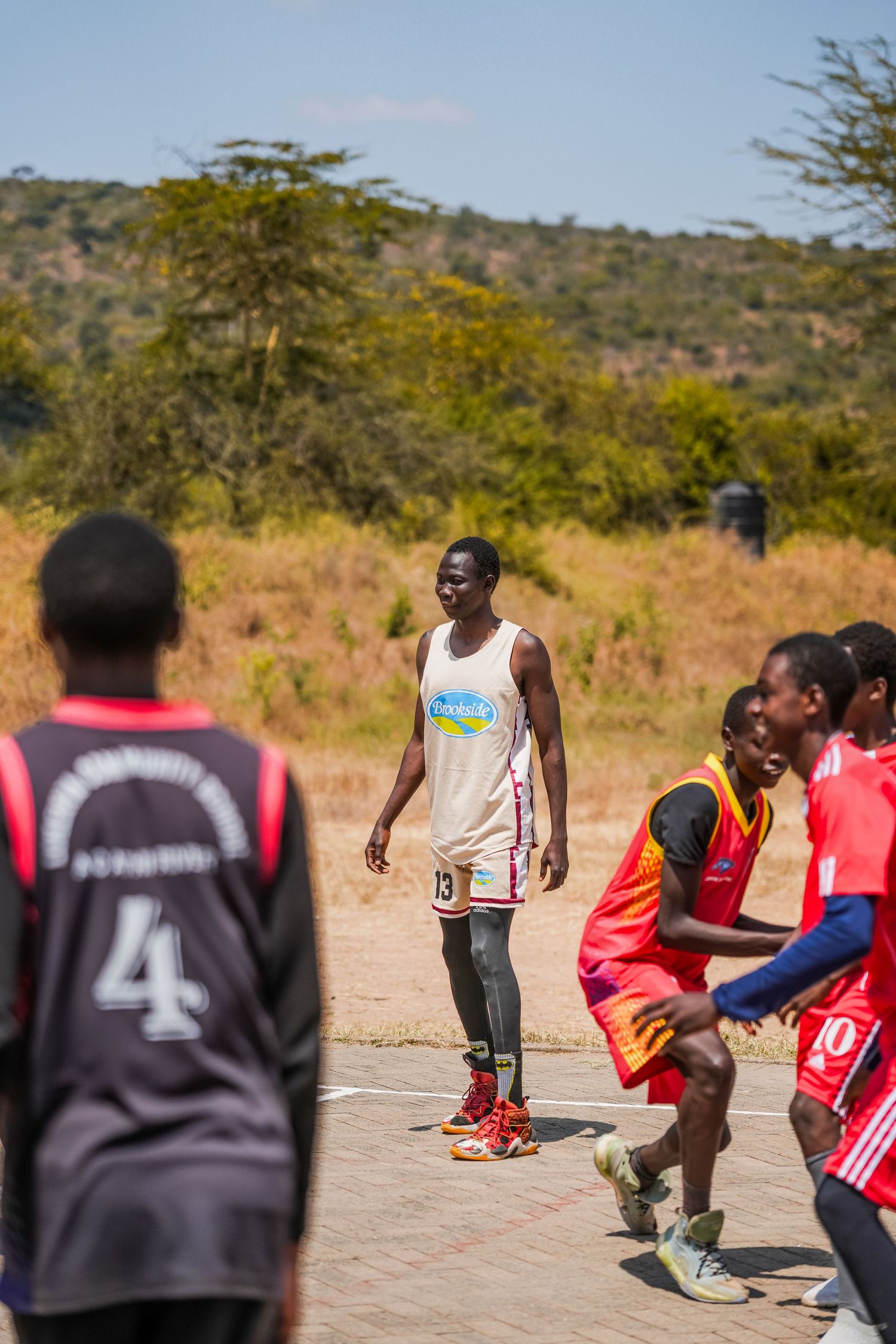 Young African men playing basketball outdoors on a court surrounded by dry savanna landscape.