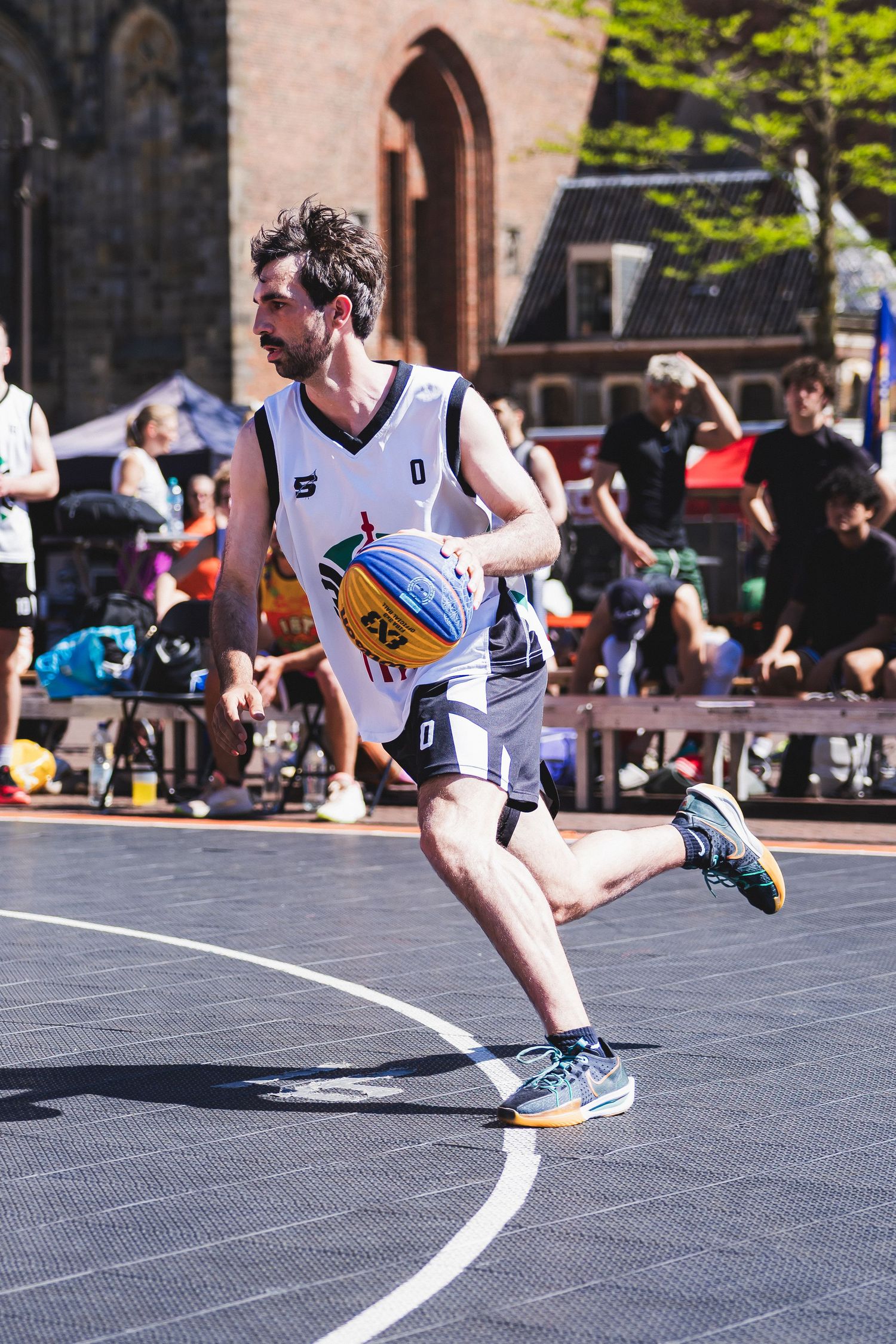 Male basketball player in white uniform dribbling ball on outdoor street court during sunny day competition.