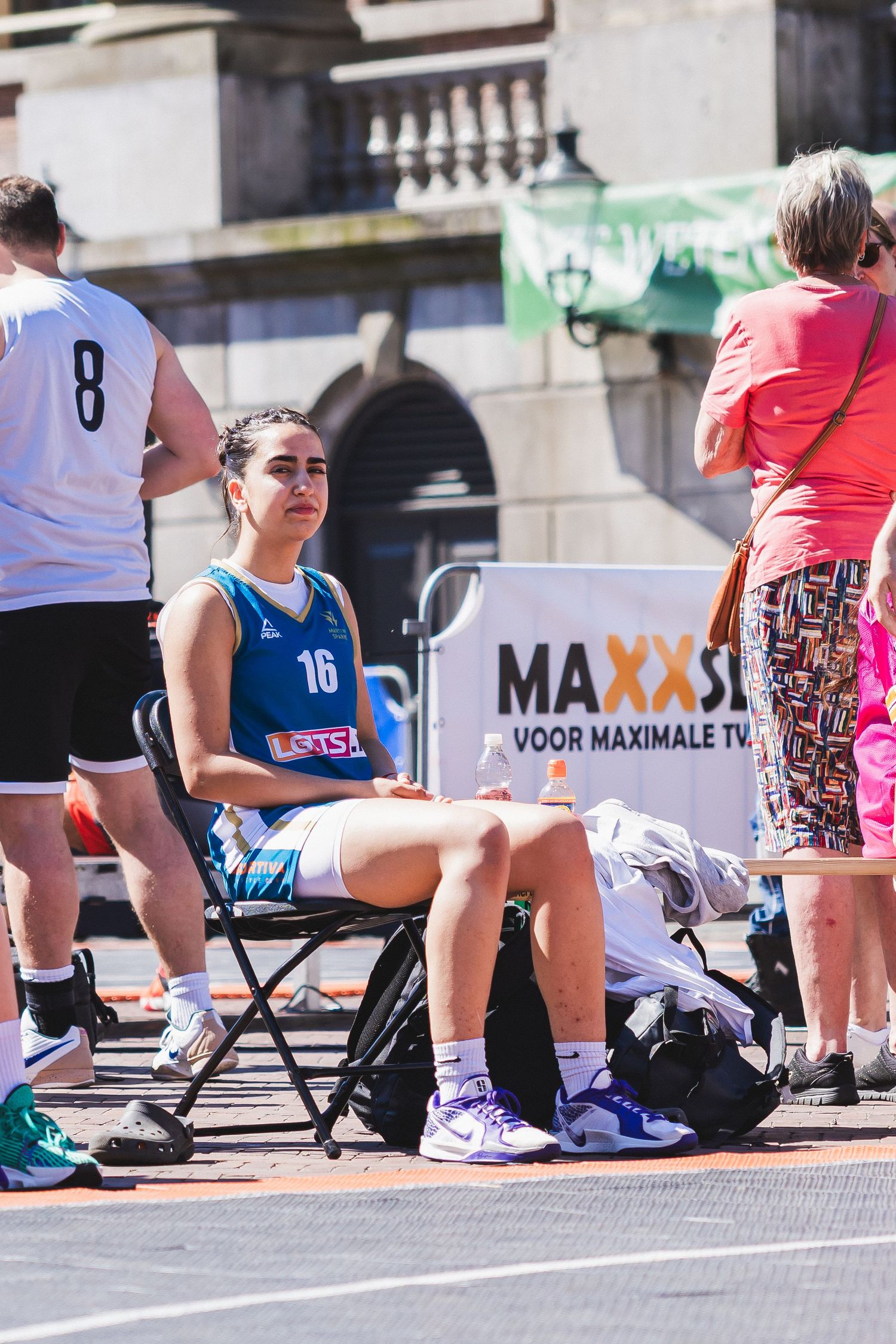 Female basketball player #16 in teal jersey sits courtside during outdoor 3x3 street basketball tournament on sunny day.