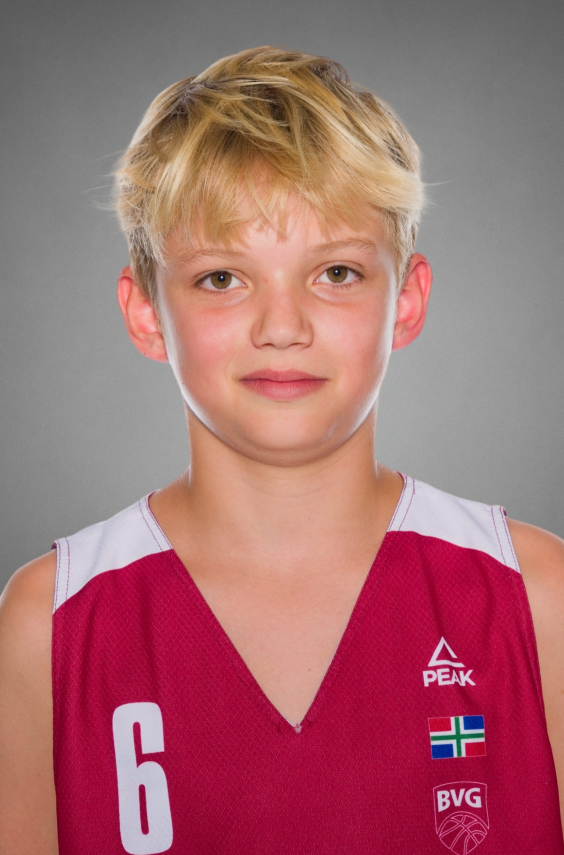 Young basketball player with blond hair wearing a red number 6 Peak jersey with BVG logo in a studio portrait against a gray background.