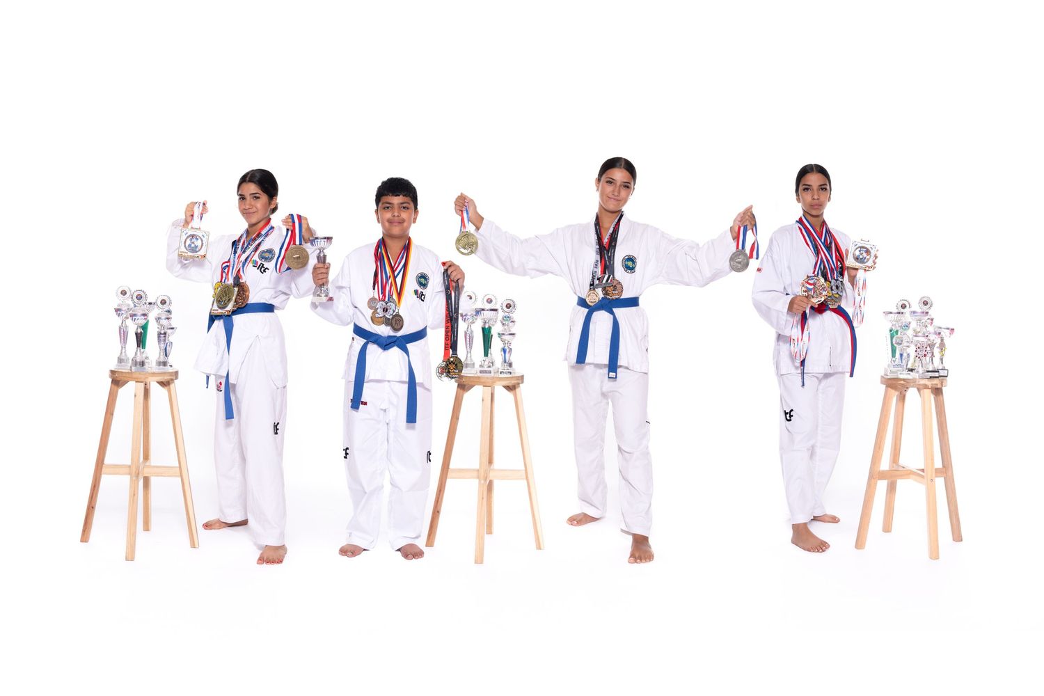 Five martial artists in white uniforms demonstrate karate poses with trophies displayed on wooden stools.