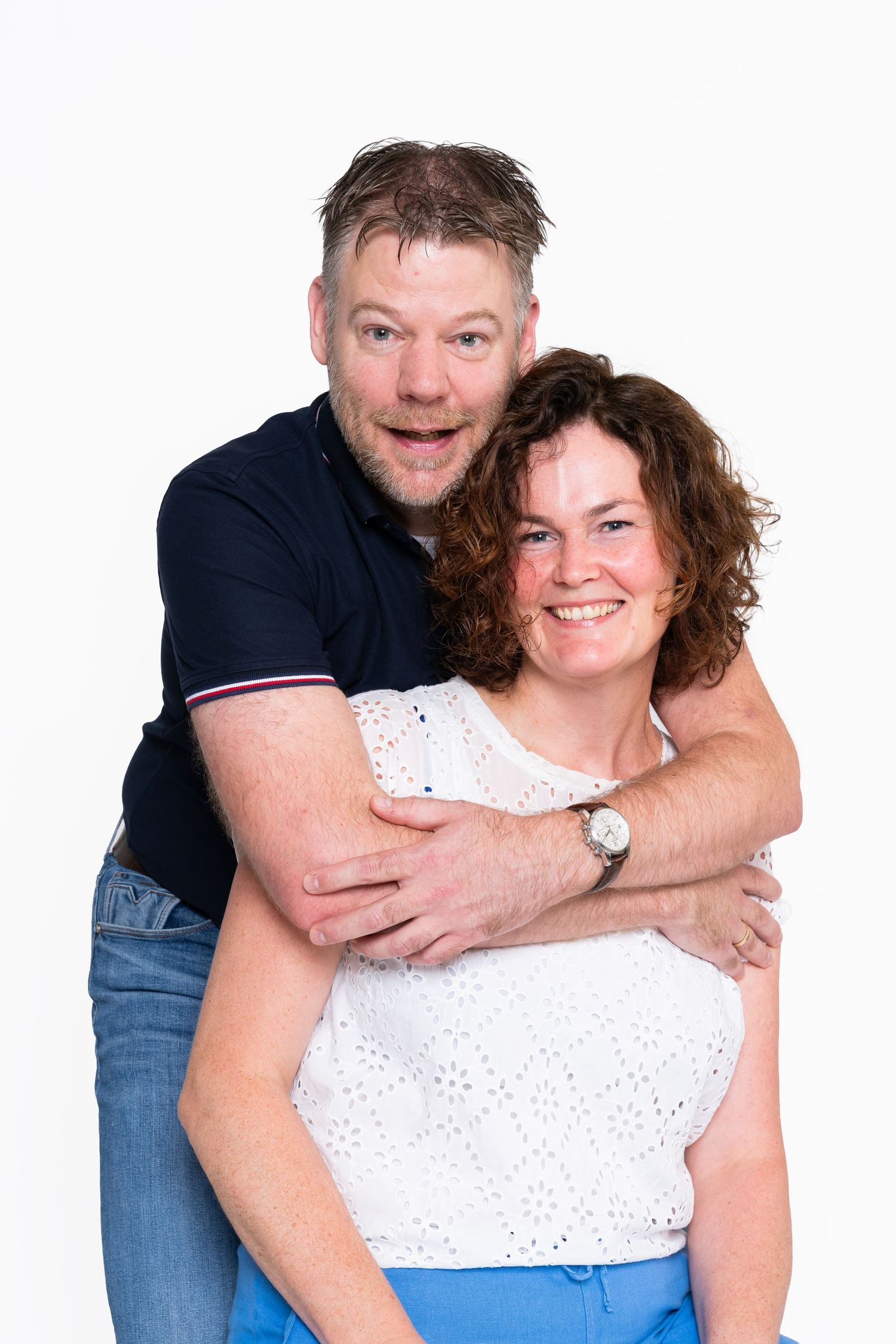 A couple embraces and smiles at the camera, wearing casual clothing against a white background.