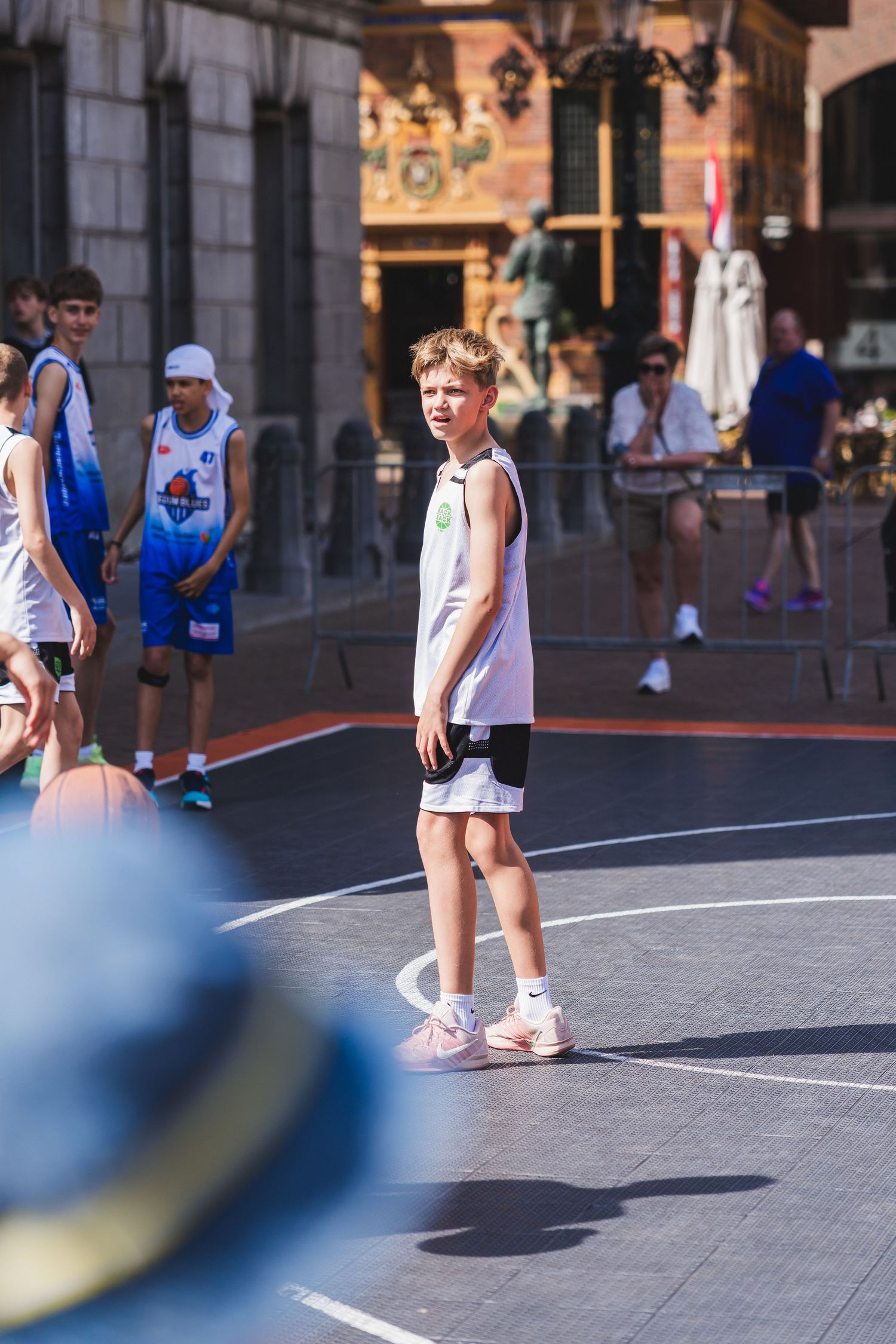 Young boy in white basketball jersey stands on outdoor urban court during a street basketball tournament on a sunny day.