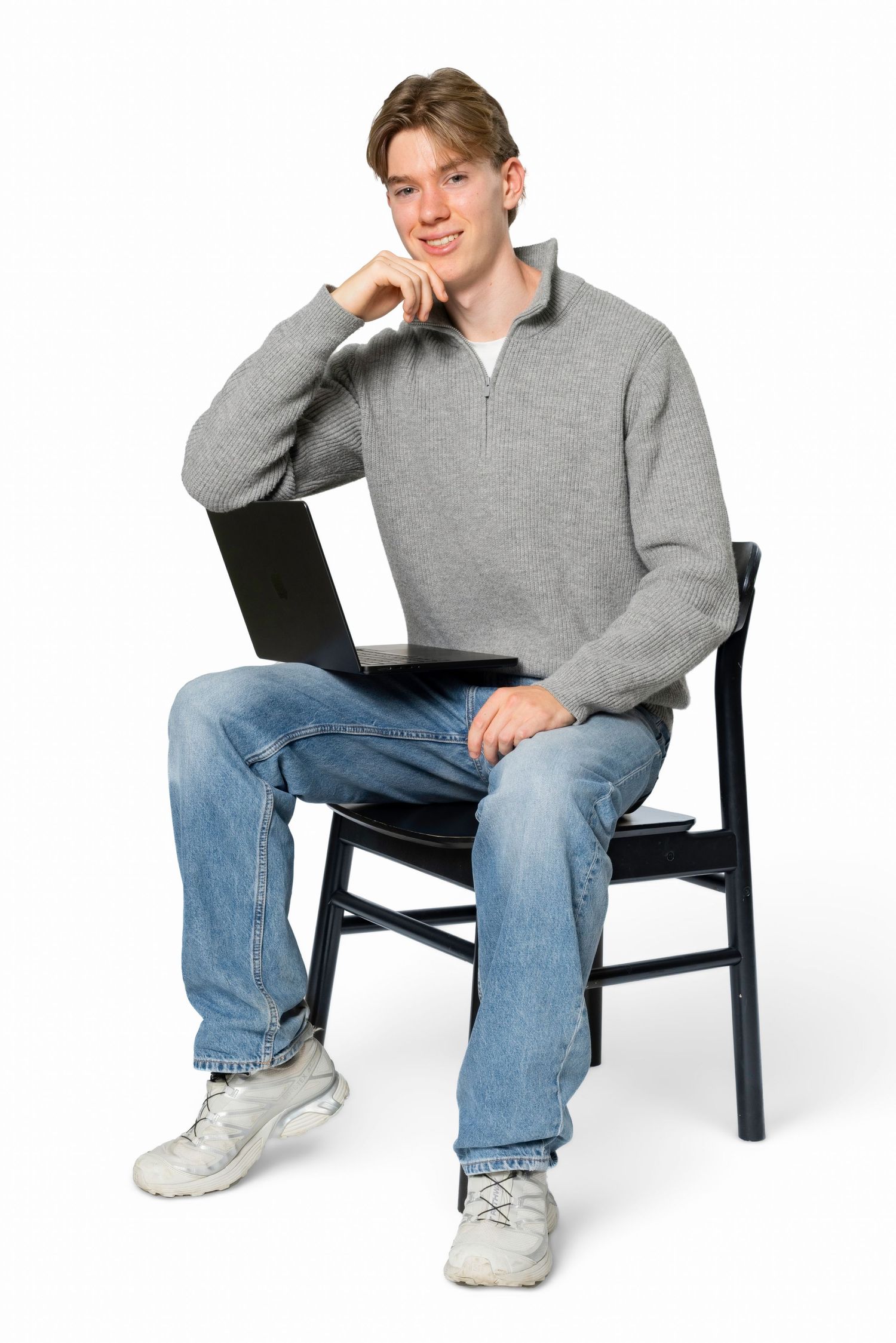 Young man in a gray quarter-zip sweater and jeans sits on a black chair, smiling with a laptop on his lap against a white background.
