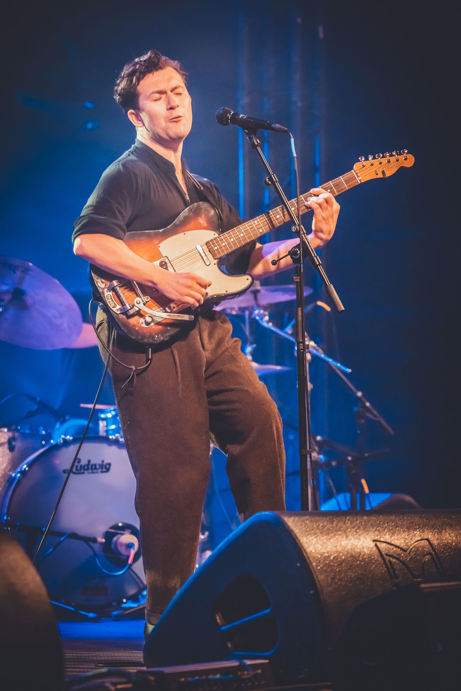 A performer plays an electric guitar on stage with dramatic blue lighting and a drum kit visible in the background.