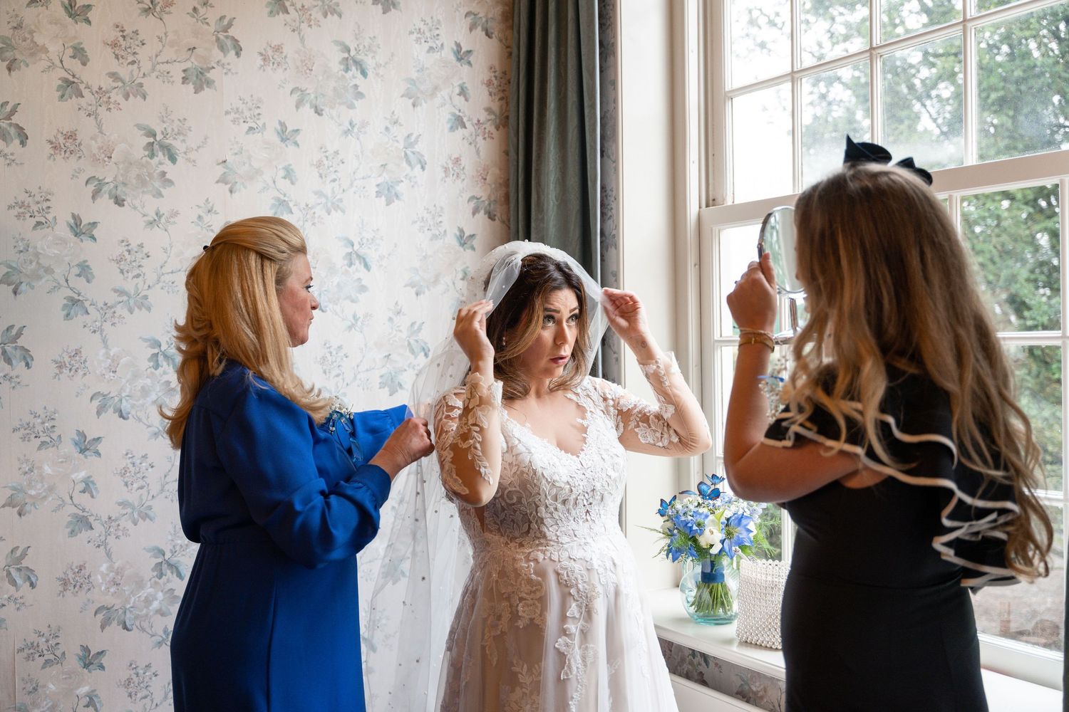 A bride in a white dress gets ready with help from two friends in a room with floral wallpaper and natural light.