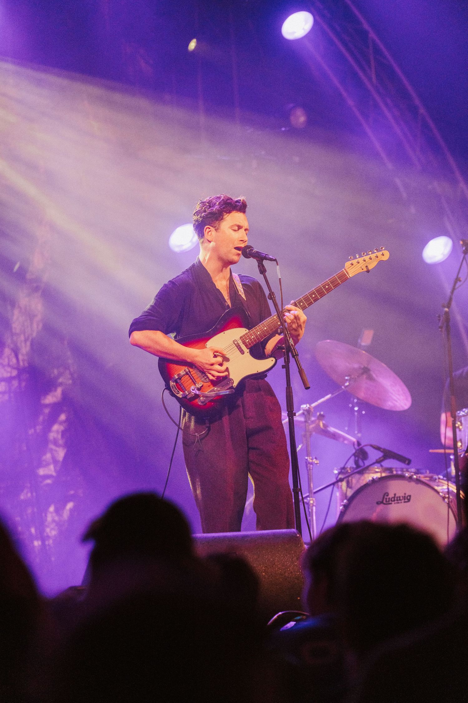 A musician performs on stage bathed in purple lighting while silhouetted audience members watch from below.