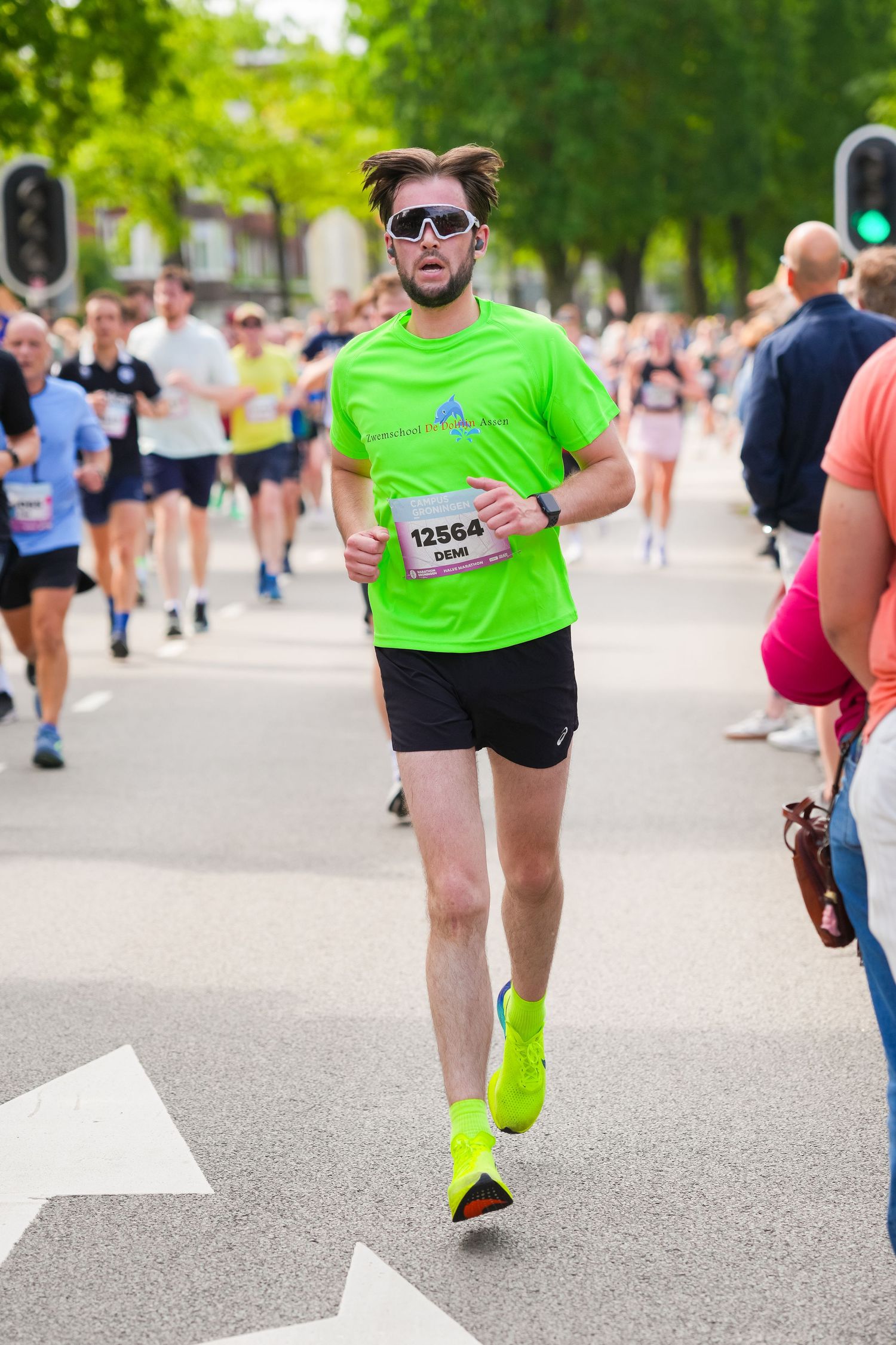 Runner #12564 in neon green shirt and yellow shoes races through a crowded marathon on a tree-lined street.