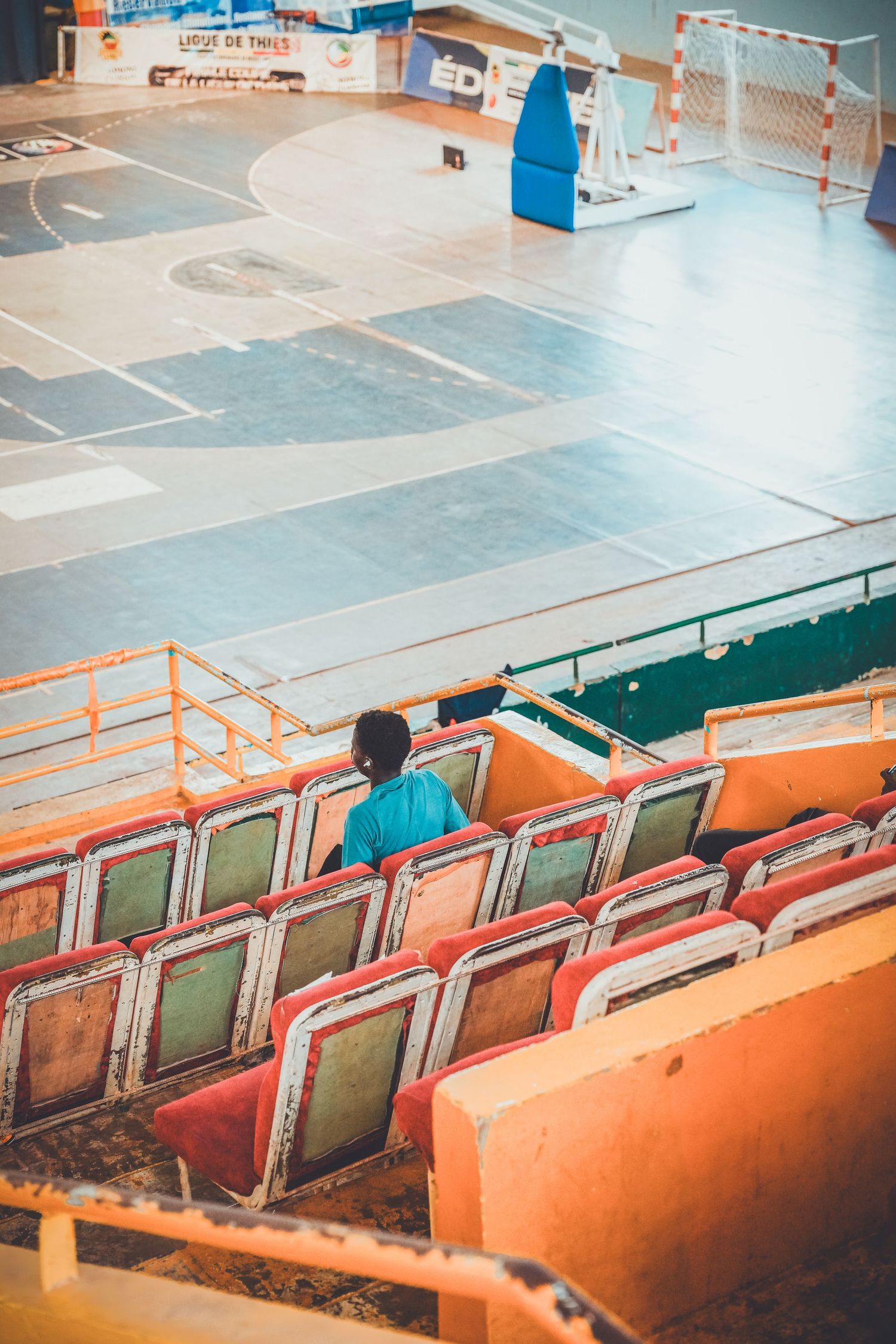 A lone child sits in worn, colorful stadium seats overlooking an empty indoor sports court with a handball goal.
