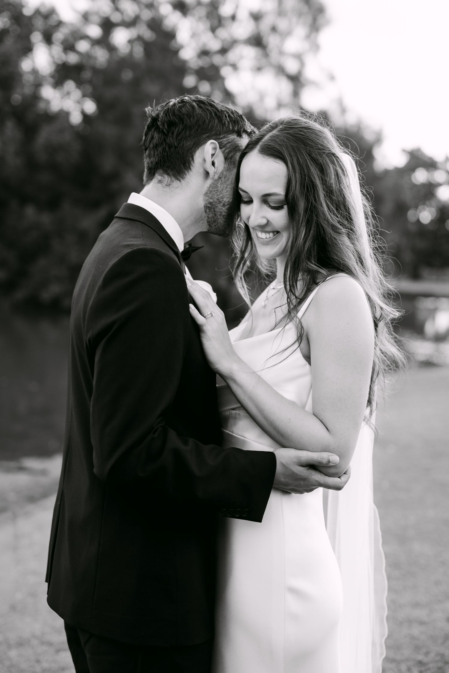 Black and white wedding photo of smiling bride in white gown embraced by groom in dark suit outdoors.