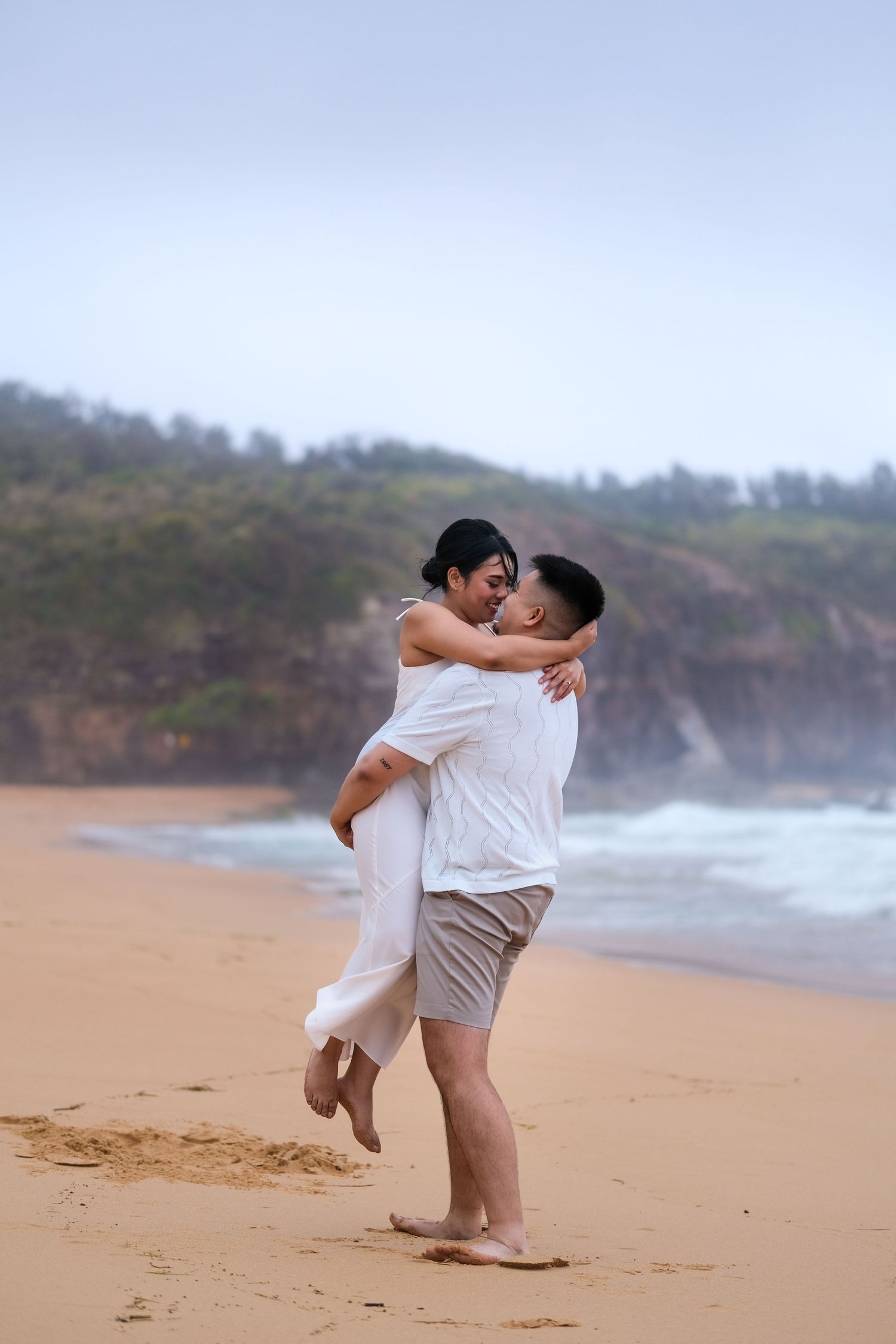 Man carrying woman in his arms while walking on a sandy beach on an overcast day.
