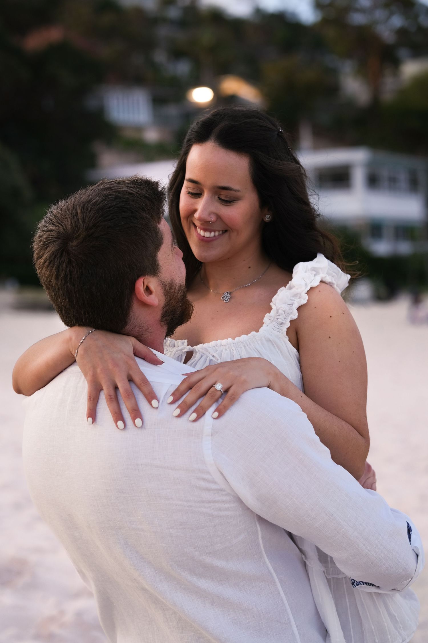 Man lifting smiling woman in white ruffled dress on sandy beach at dusk, engagement ring visible on her hand.