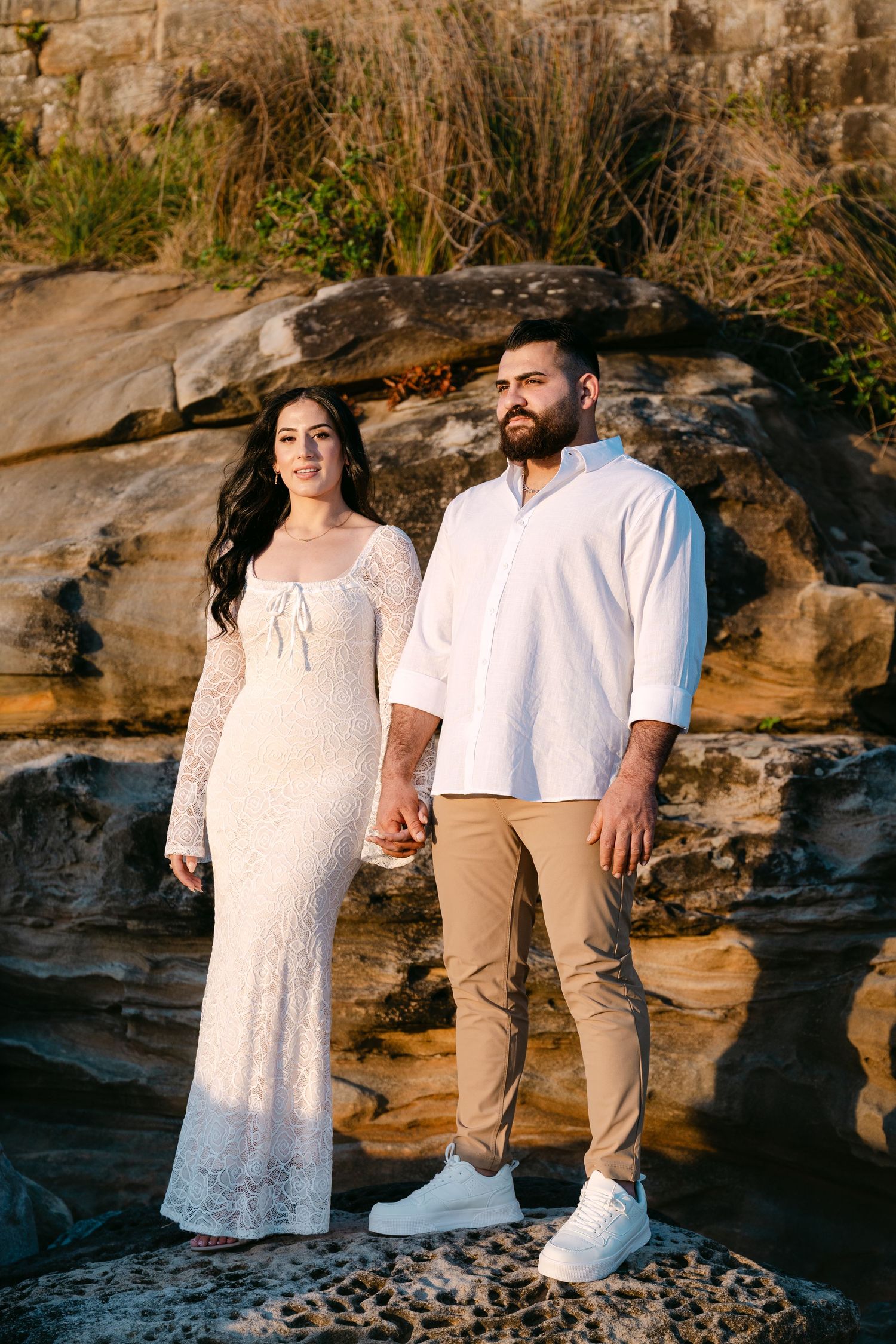 Couple standing on rocks at golden hour, woman in white lace dress and man in white shirt holding hands.