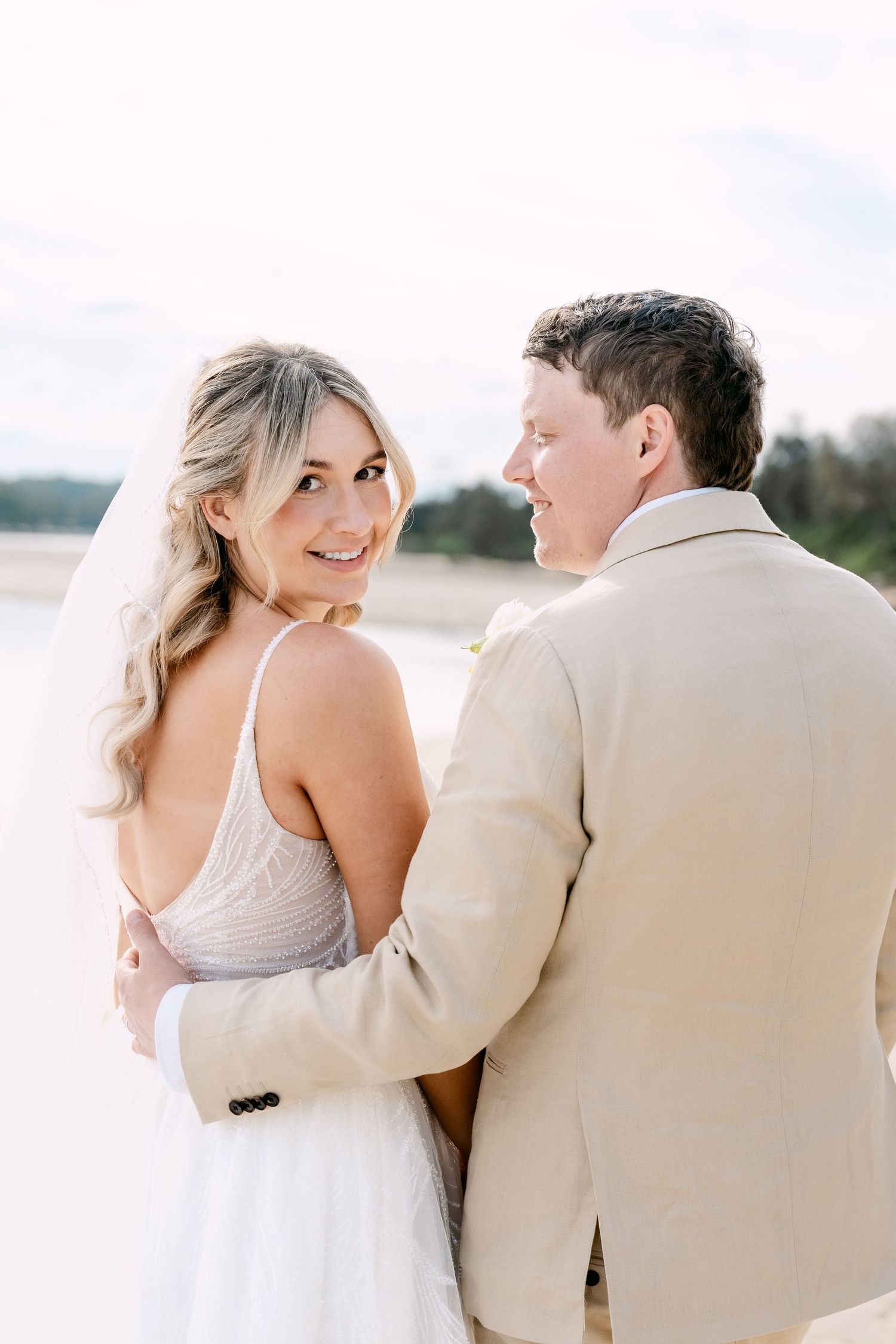 Bride in white gown and groom in tan suit embrace on sandy beach at outdoor wedding.