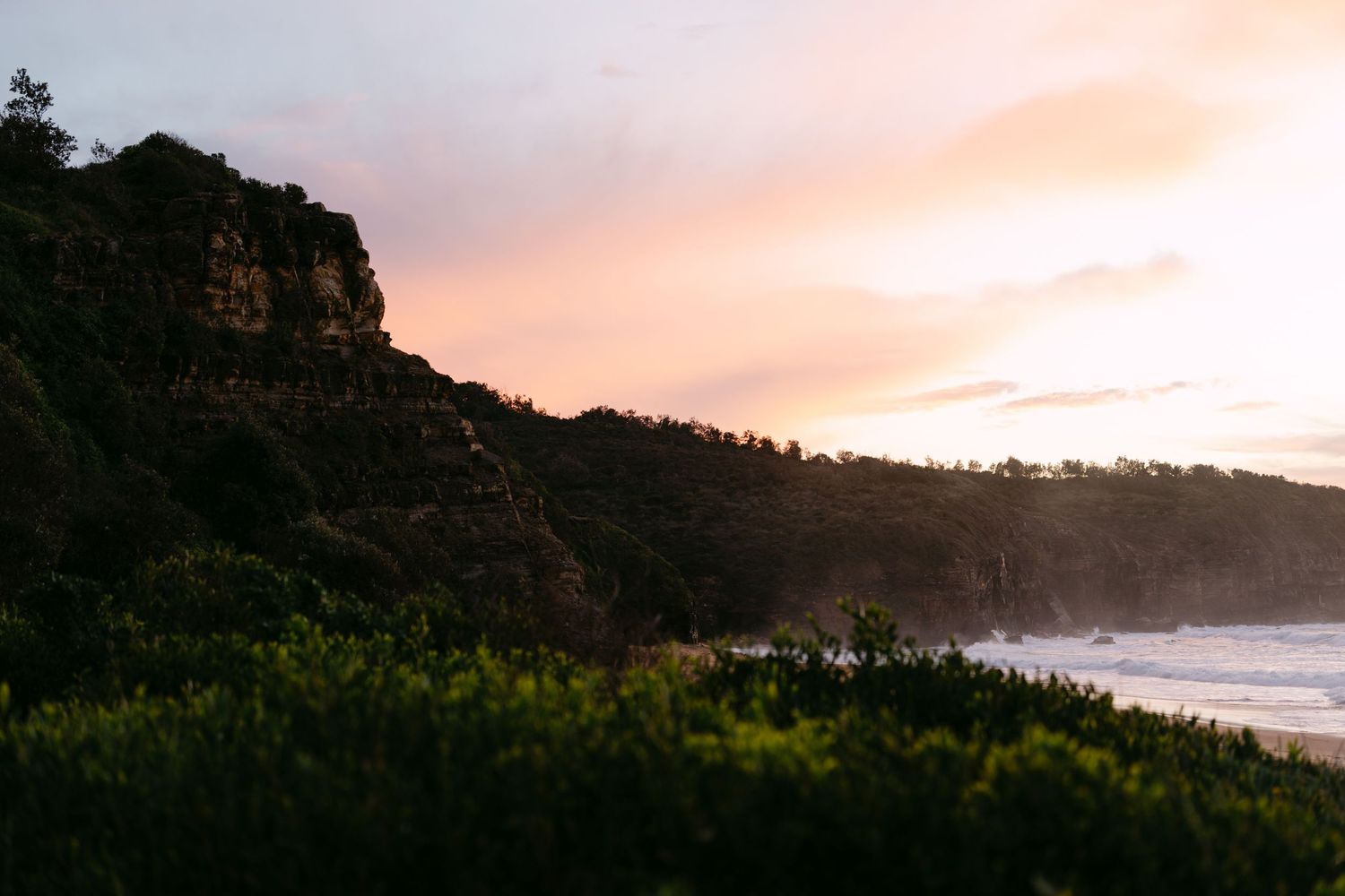 Stunning coastal sunset with rocky cliffs, lush green vegetation, and ocean waves under a pink and orange sky.