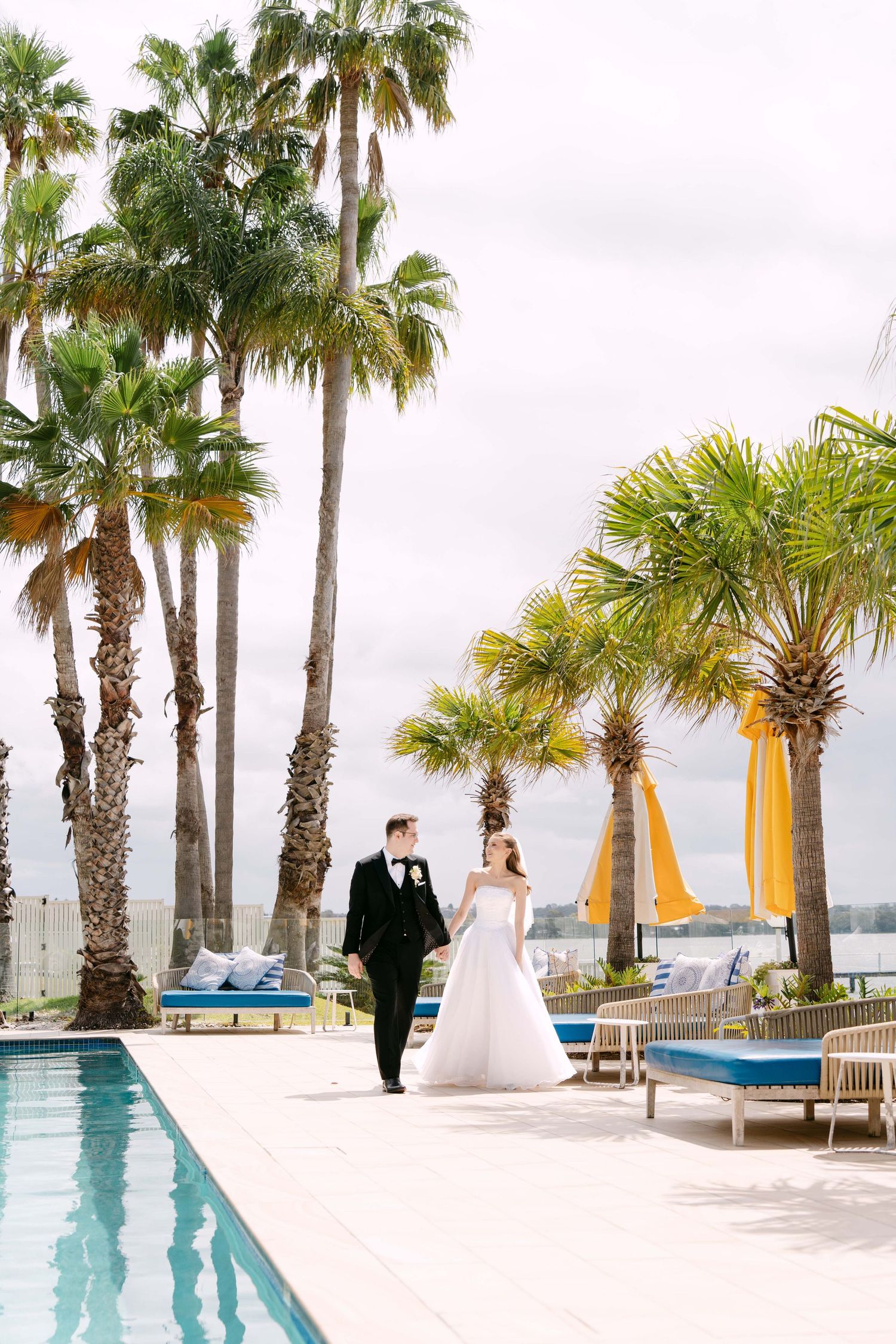 Bride in a white gown and groom in a black tuxedo walk hand in hand beside a pool lined with palm trees and yellow umbrellas.