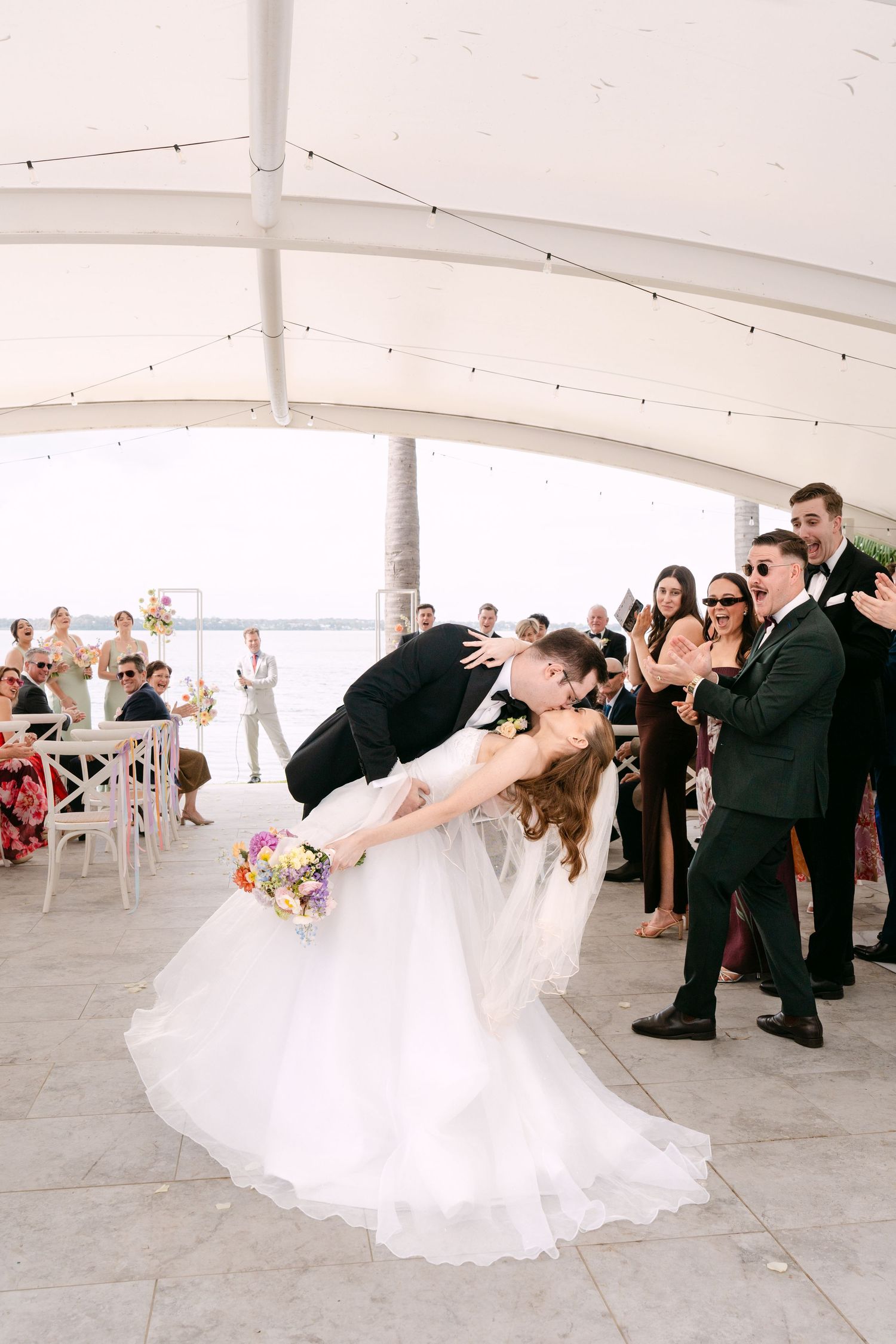Groom dips bride in a romantic kiss under a white tent as wedding guests cheer and clap at a waterfront outdoor ceremony.