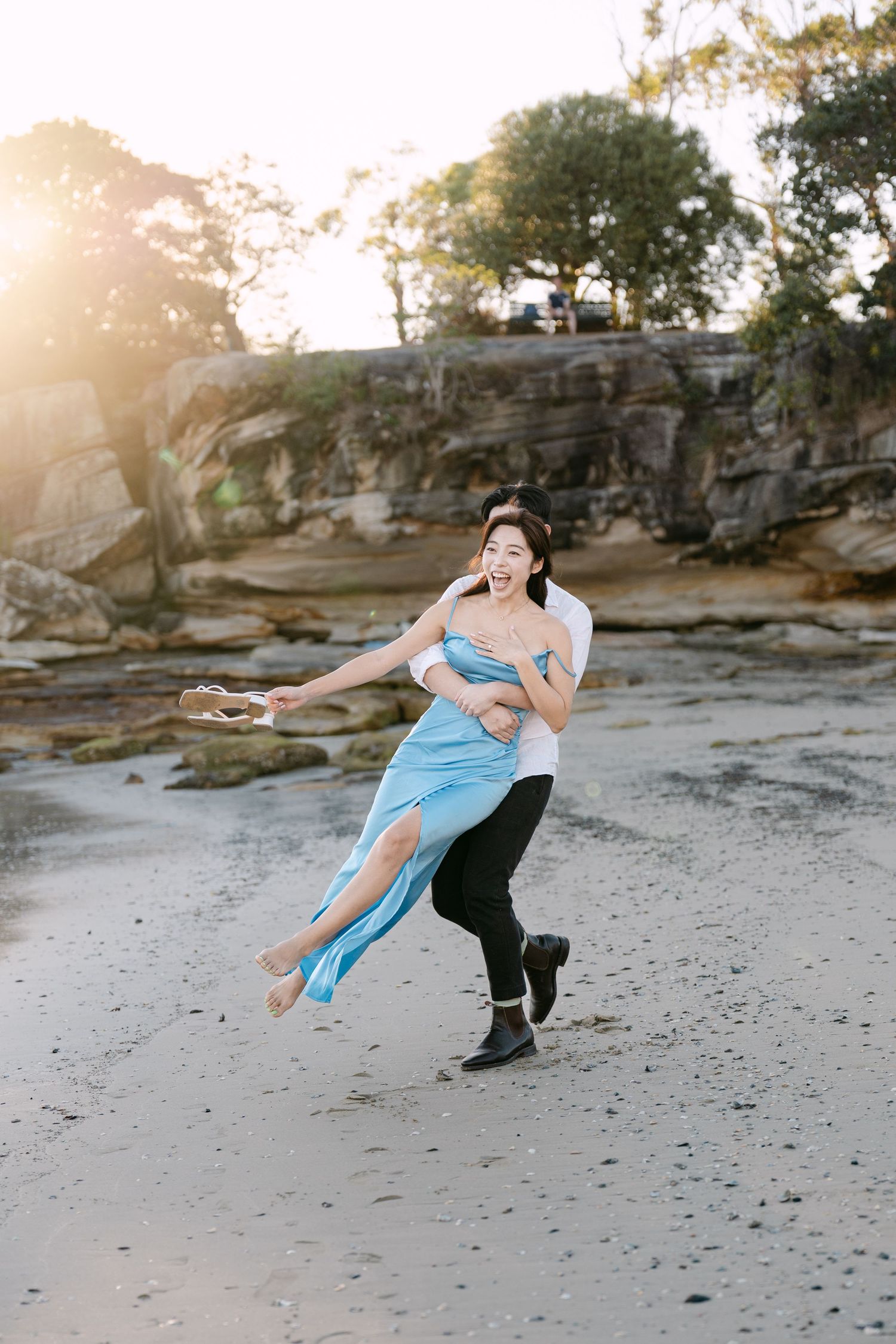 Couple joyfully spinning on a sandy beach at golden hour, woman in blue dress laughing as man lifts her.