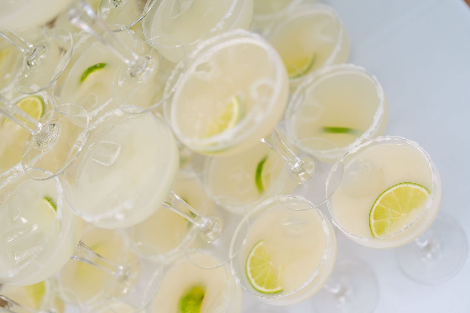 Overhead view of many stemmed glasses filled with a pale cocktail and garnished with lime wedges and slices, arranged closely together.