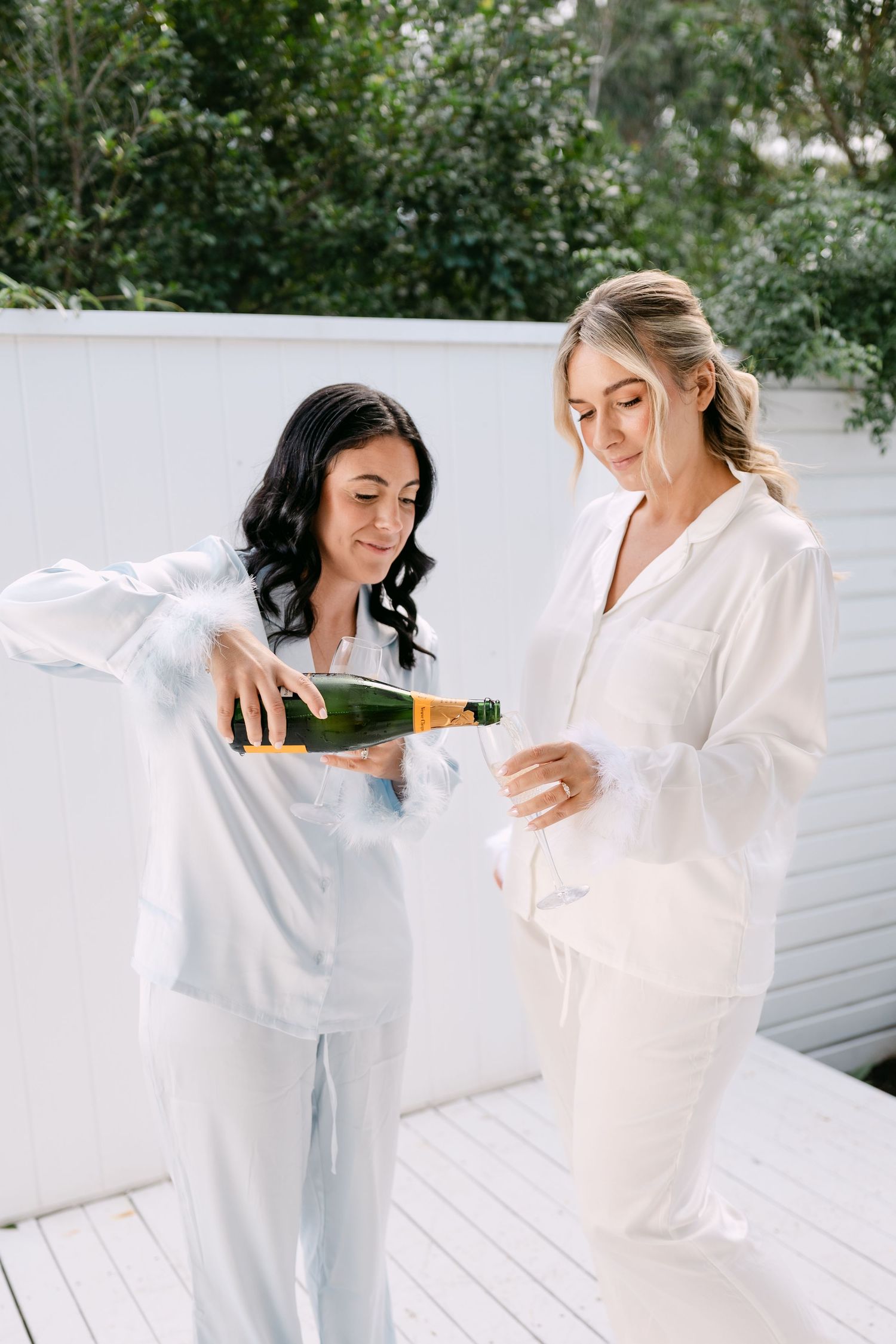 Two women in white satin pajamas pouring champagne on a sunny outdoor deck surrounded by greenery.