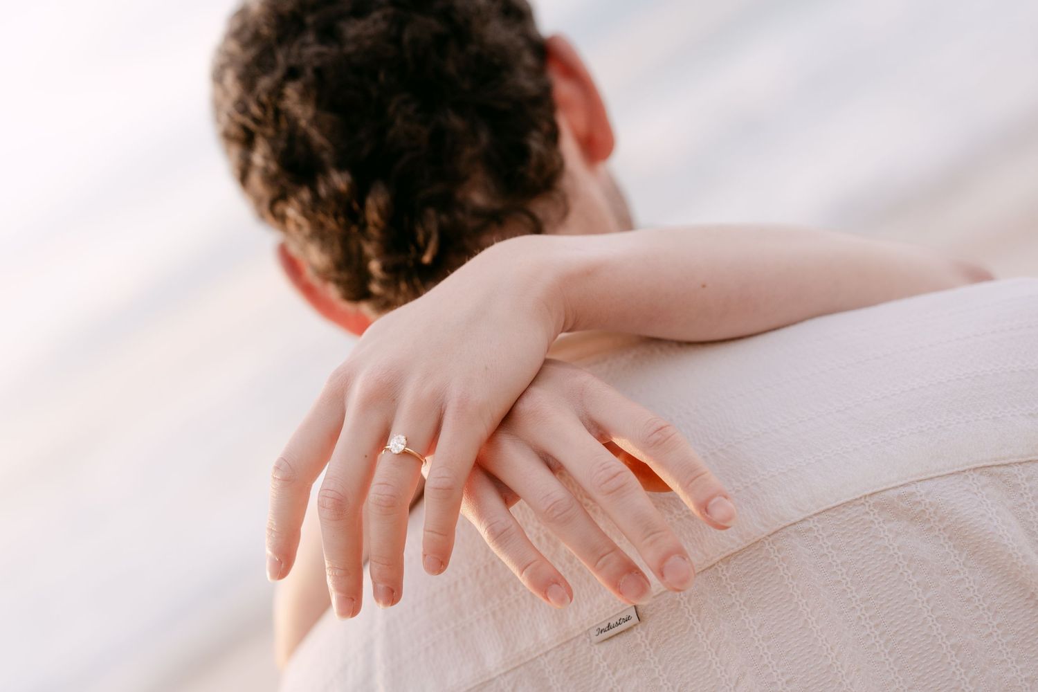 Detail shot of diamond engagement ring on woman's hand resting on groom's shoulder at beach.