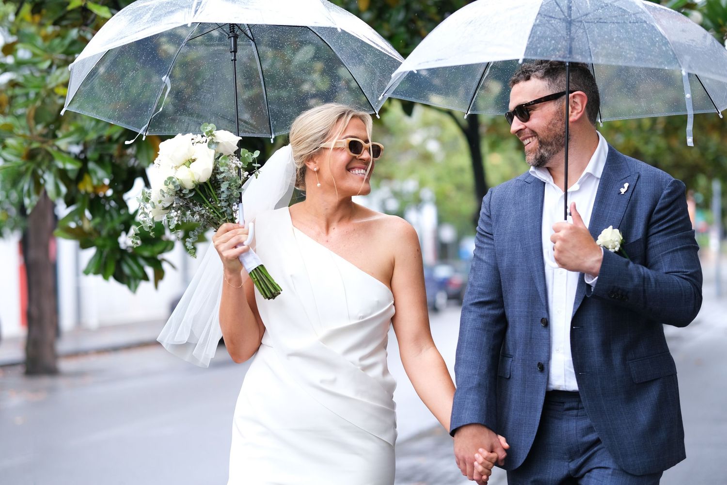 Smiling bride and groom walk hand in hand under clear umbrellas on a rainy city street.