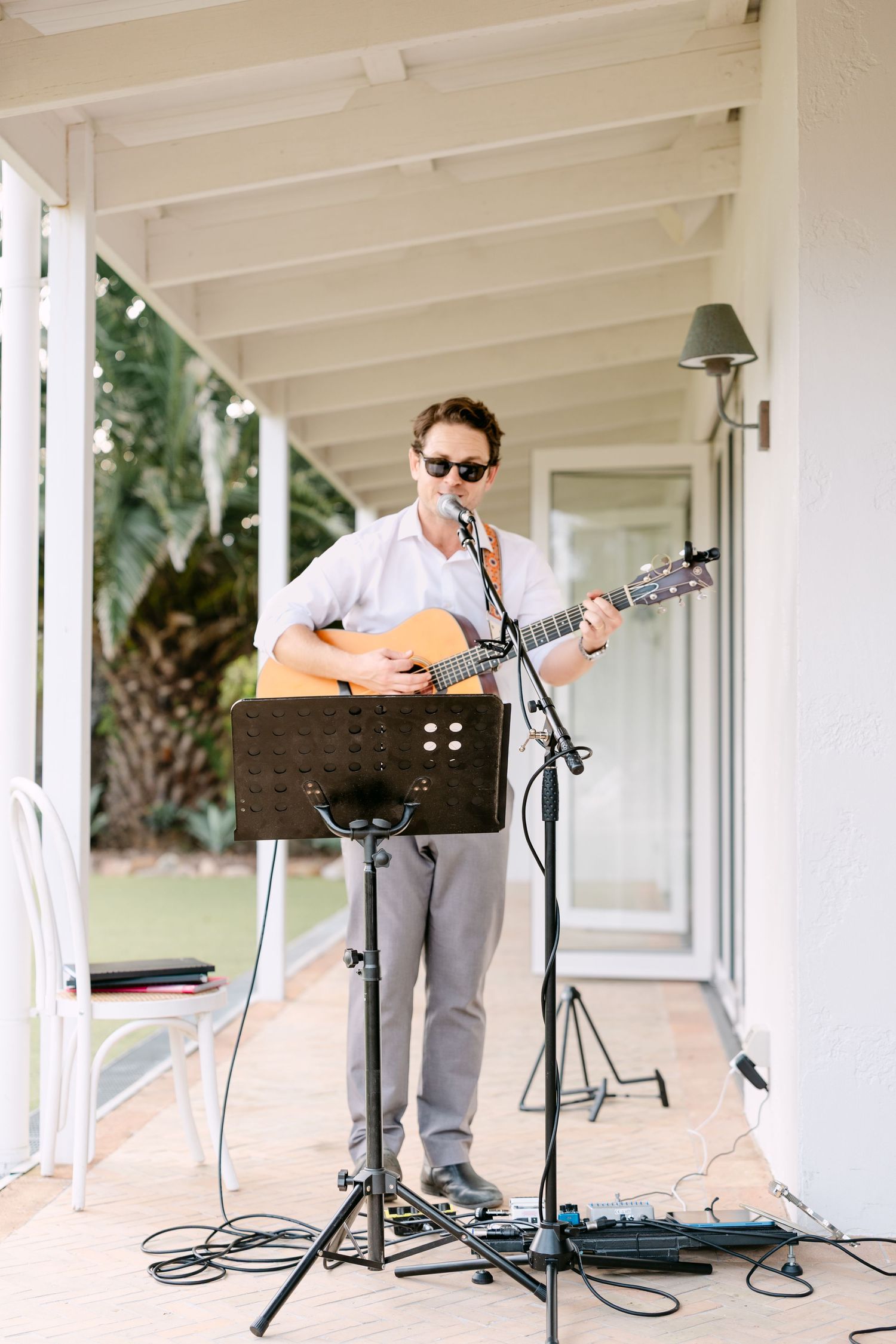 Musician in sunglasses plays acoustic guitar and sings into a microphone on a veranda at a wedding.