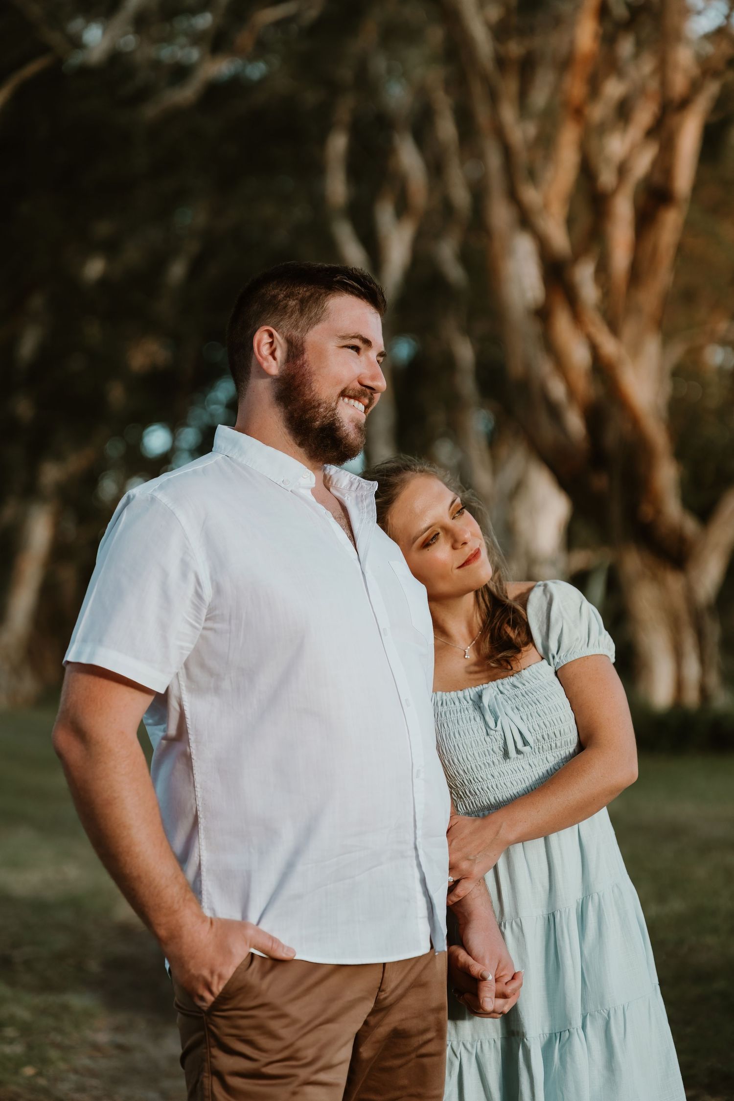 Bearded man in white shirt with woman in blue dress leaning on him in a sunlit park.