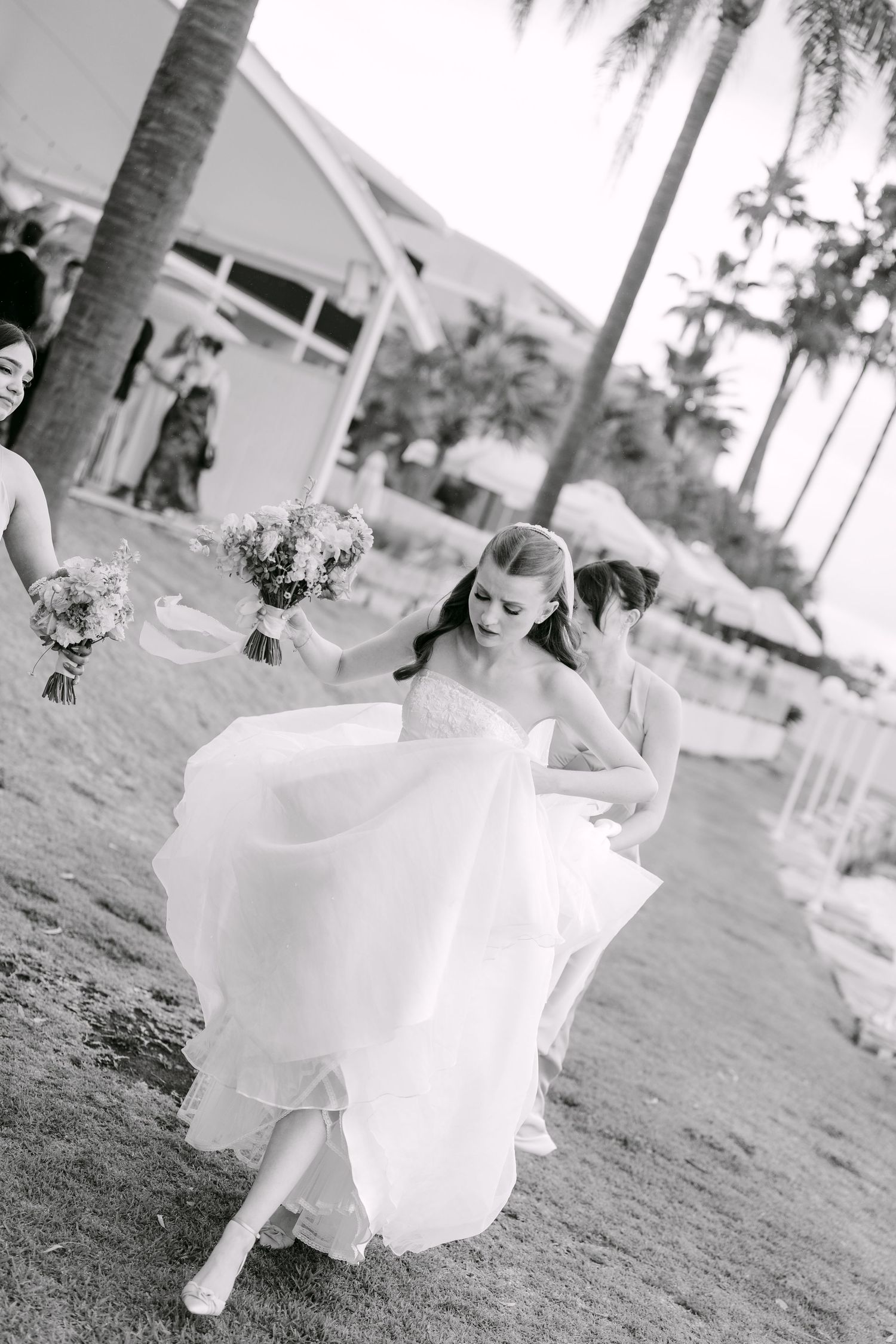 Black and white photo of a bride in a flowing tulle gown lifting her skirt as she walks past bridesmaids holding bouquets near palm trees.