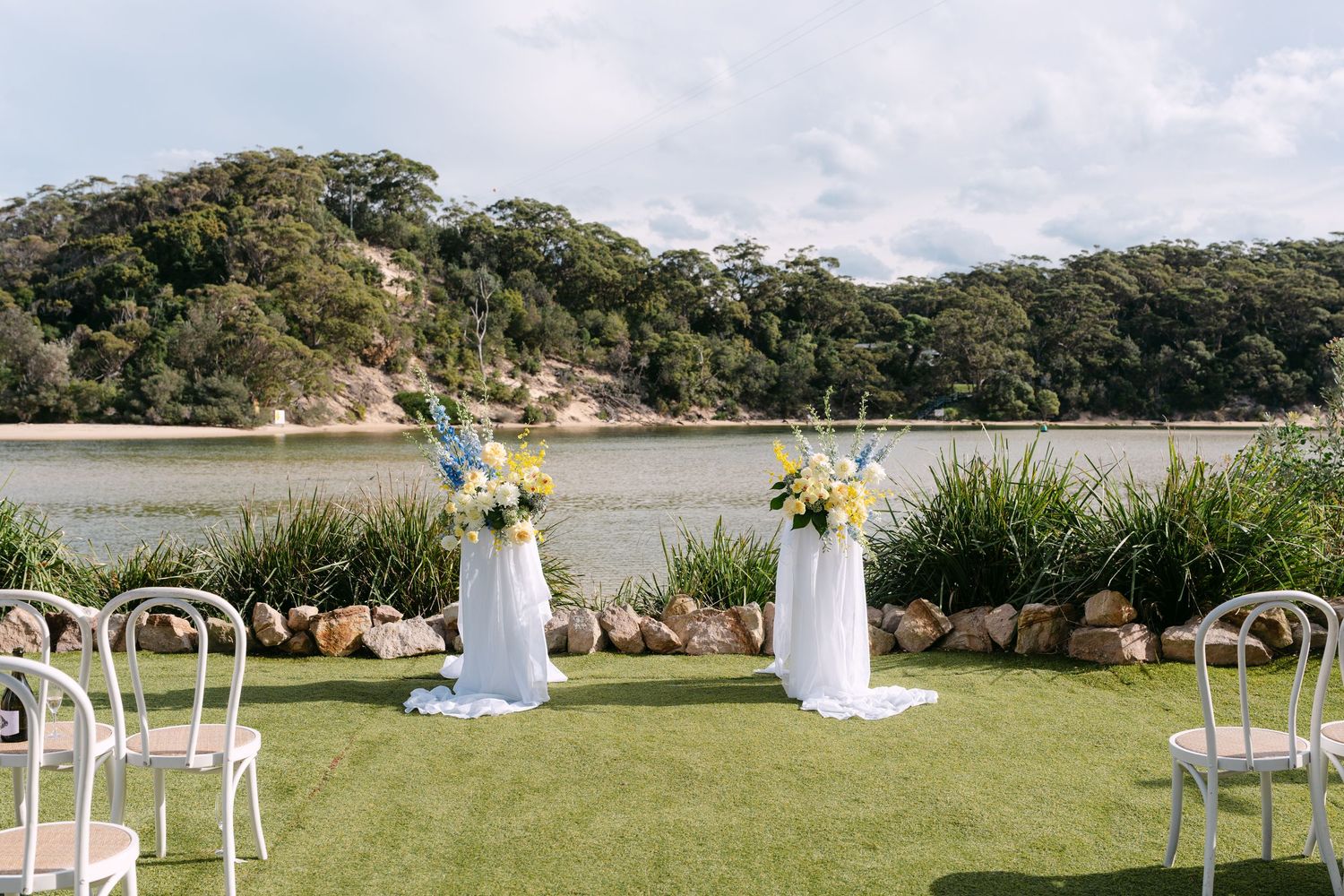 Elegant outdoor waterfront wedding ceremony setup with floral arrangements, white chairs, and scenic lake backdrop.