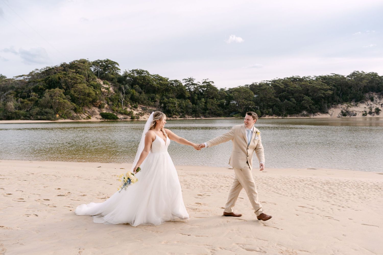 Bride in flowing white gown and groom in tan suit stroll hand in hand along a sunlit beach.