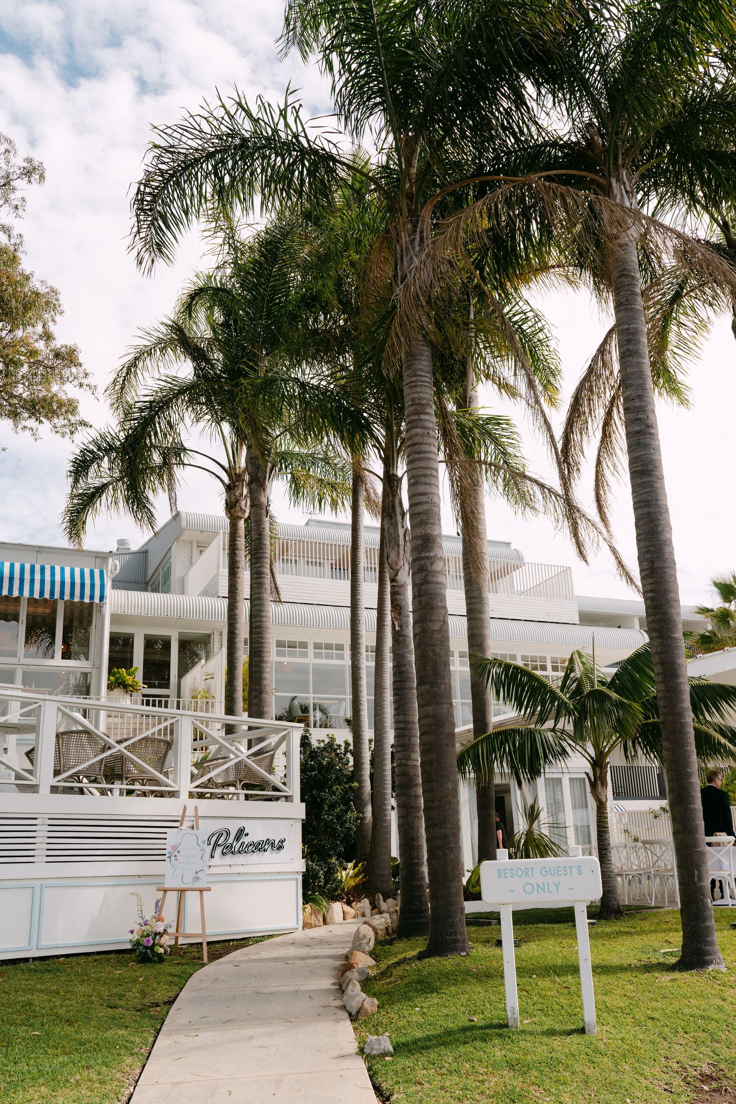 Tall palm trees line a curved concrete path leading to a white coastal hotel with a striped blue awning and signs on the lawn.