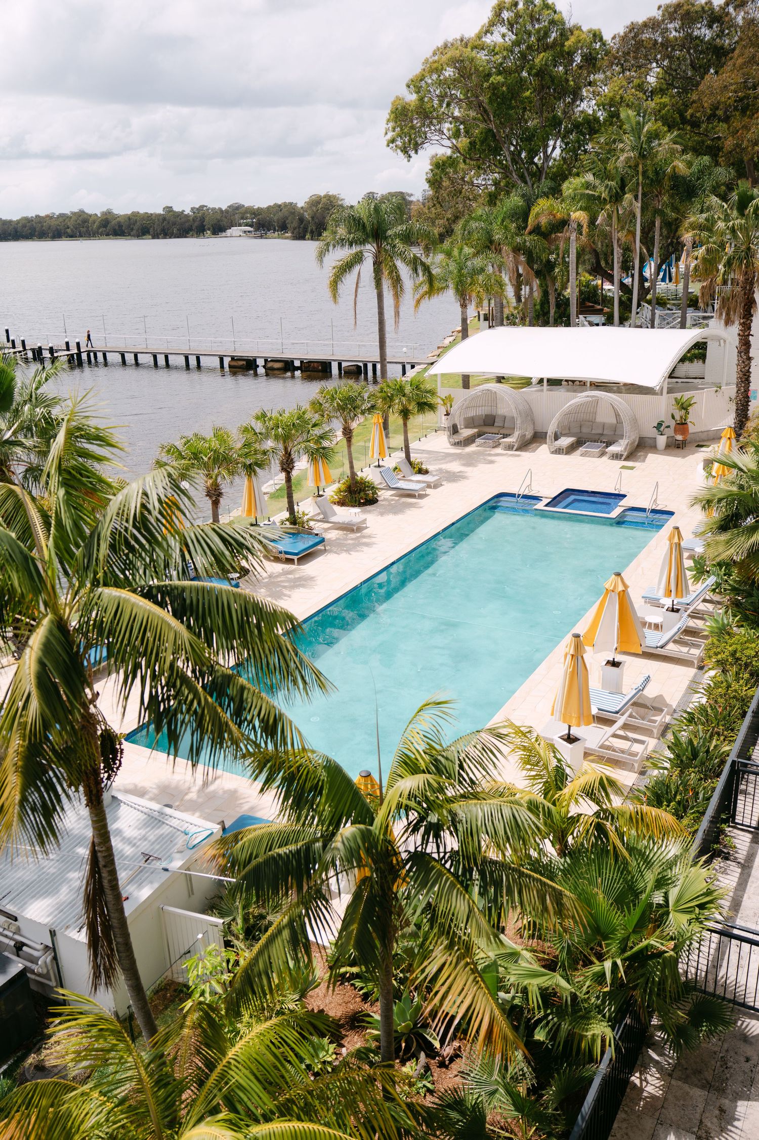 Aerial view of a resort swimming pool surrounded by palm trees, yellow umbrellas and loungers beside a calm waterfront with a jetty.
