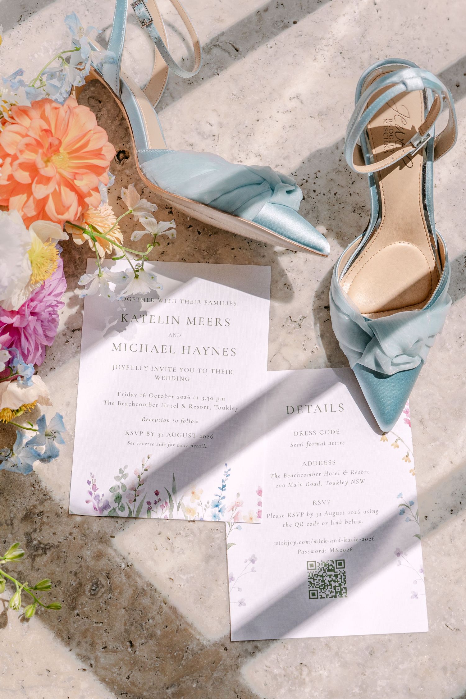 Light blue satin ankle-strap wedding heels with bow details beside a floral bouquet and printed wedding invitation and details cards.