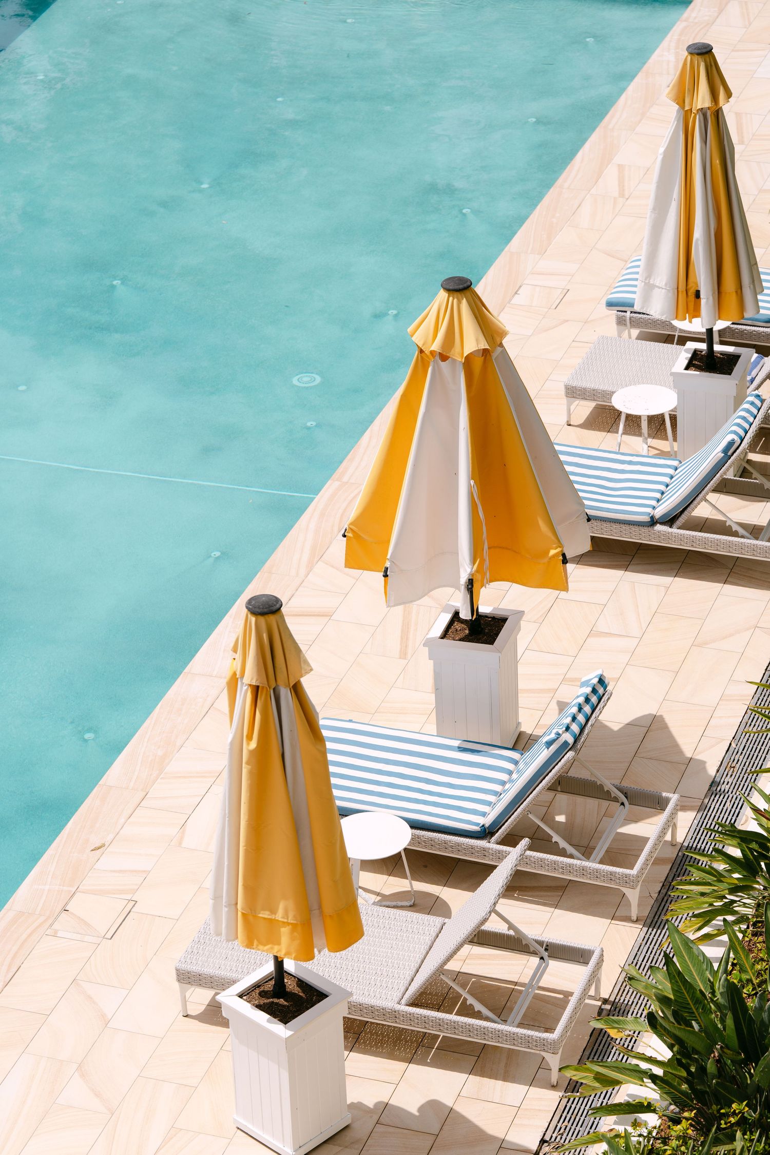 Yellow and white striped umbrellas and blue striped lounge chairs on a sunny tiled pool deck beside clear turquoise water.