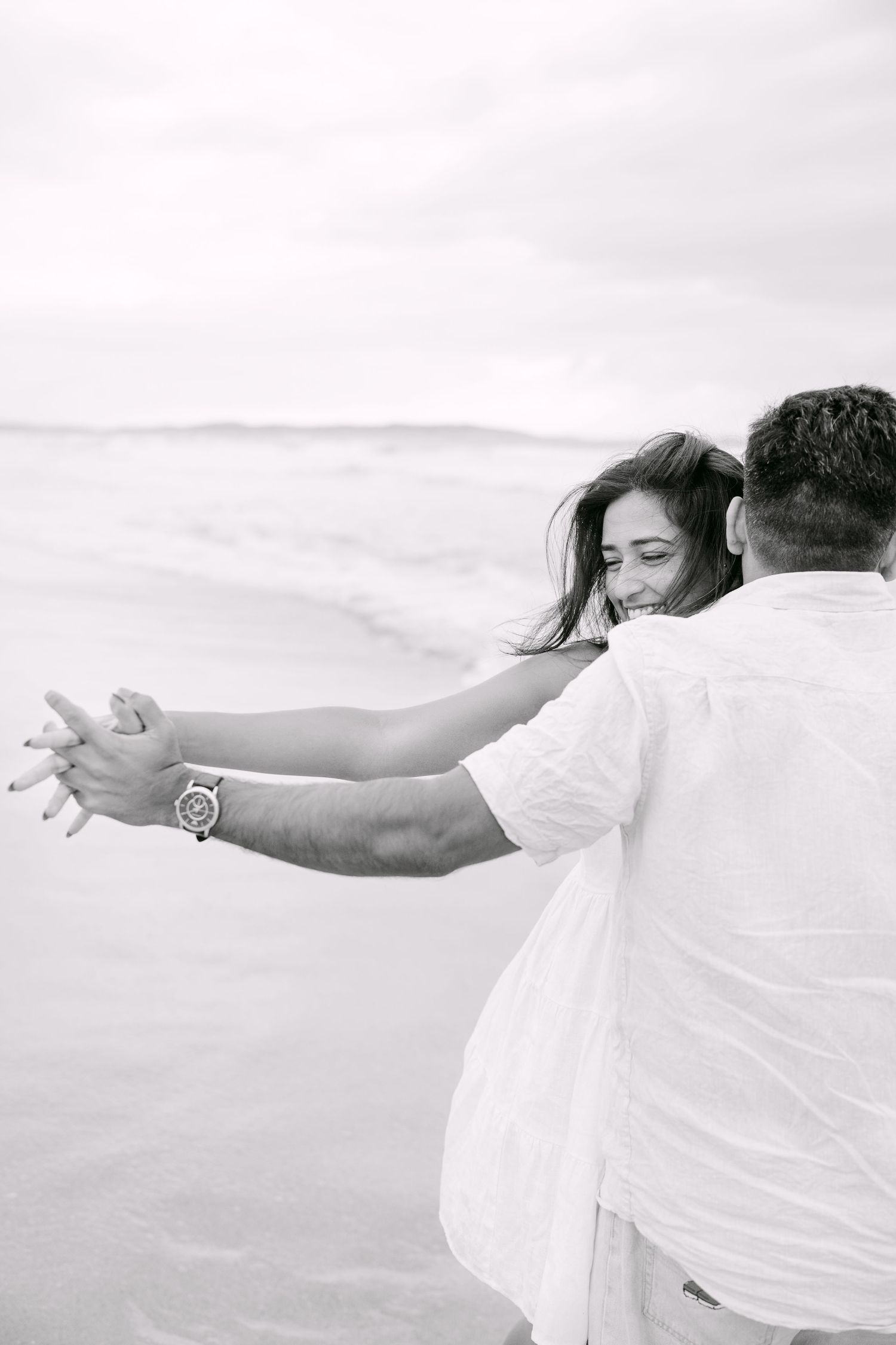A black and white photo of a couple holding hands and dancing joyfully on a beach.
