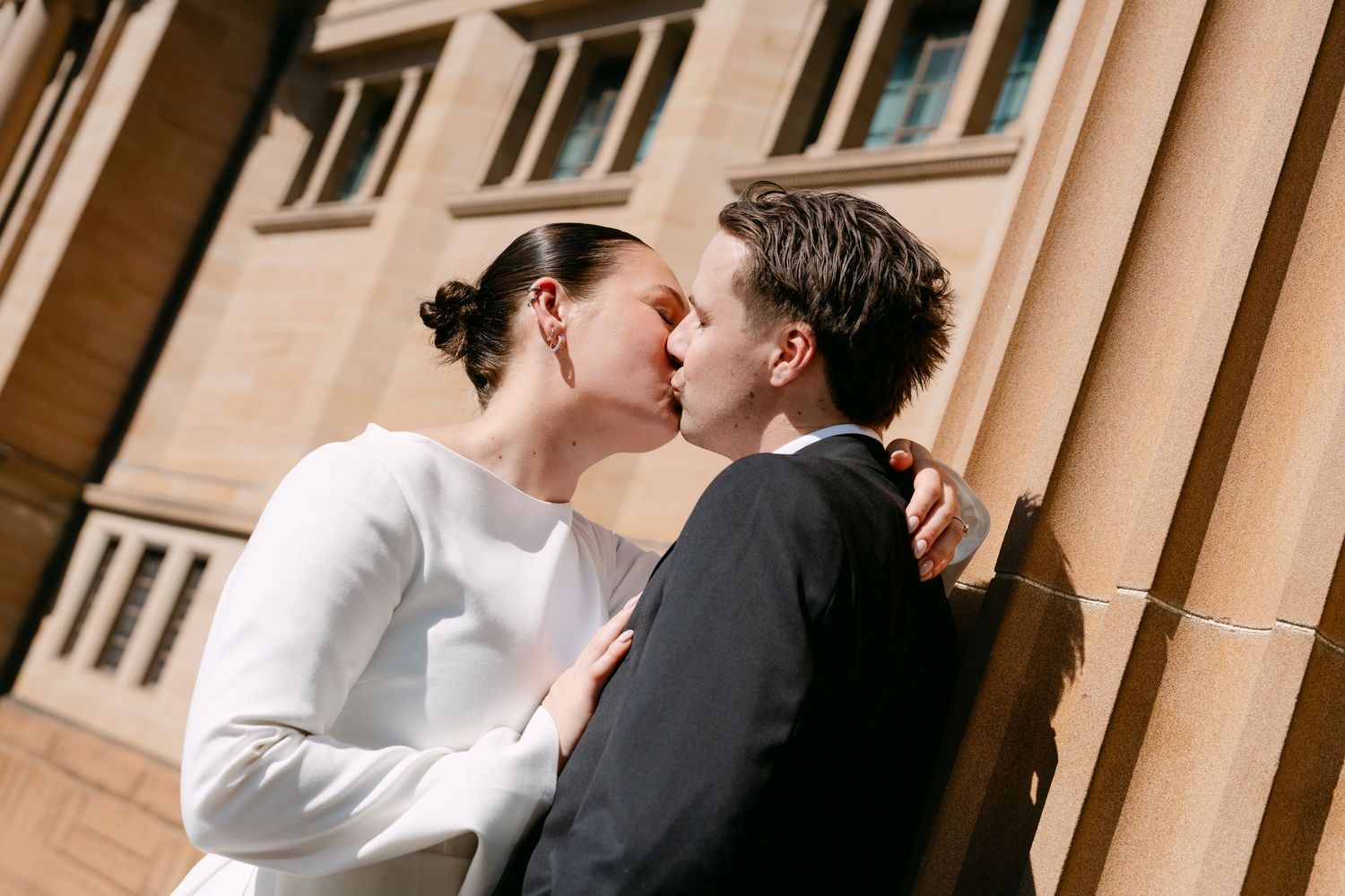 Couple sharing a romantic kiss against a sunlit stone building, bride in white dress and groom in dark suit.