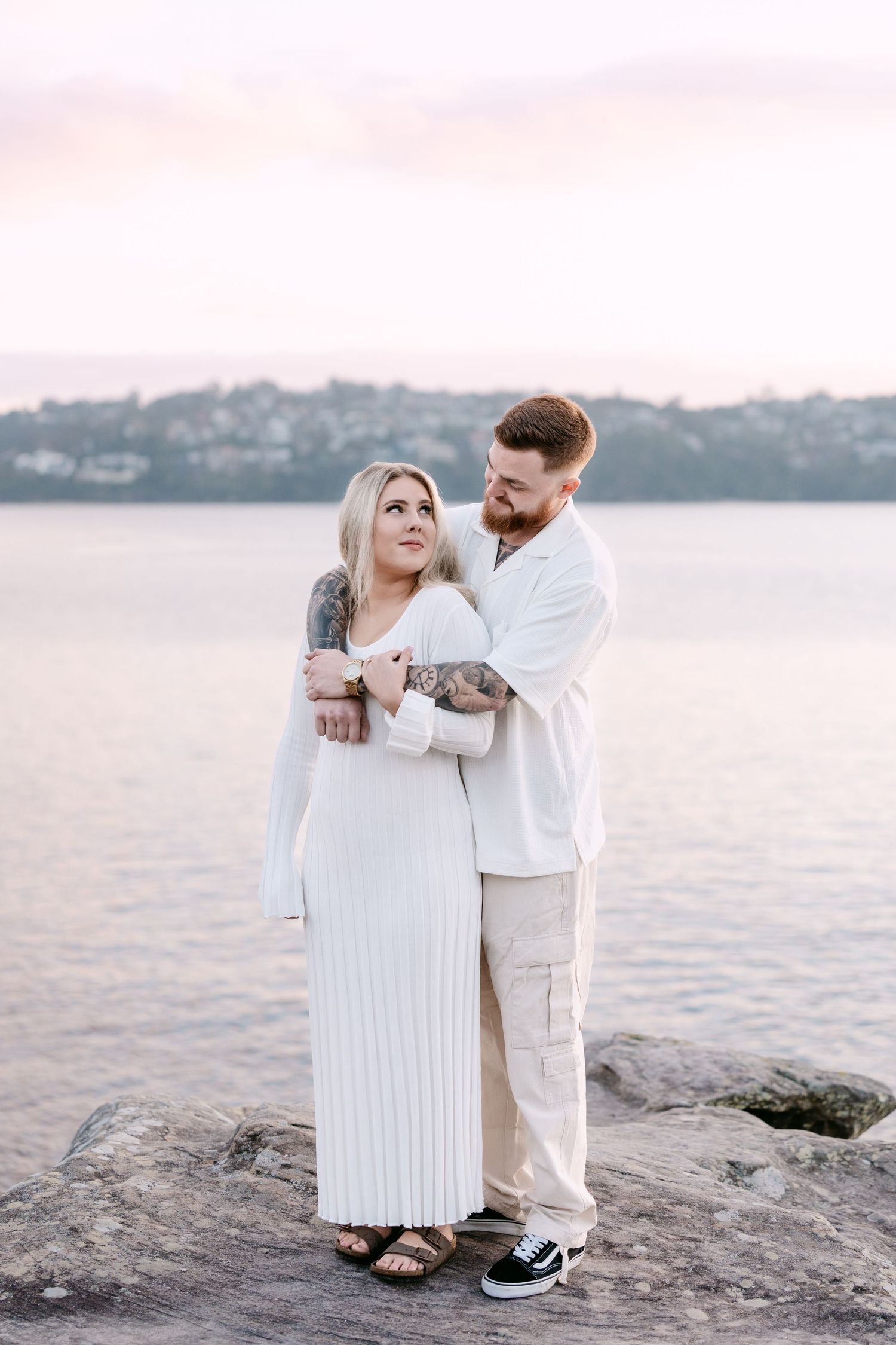 A couple in white outfits embracing by the waterfront at sunset, the man hugging the woman from behind on rocky shore.