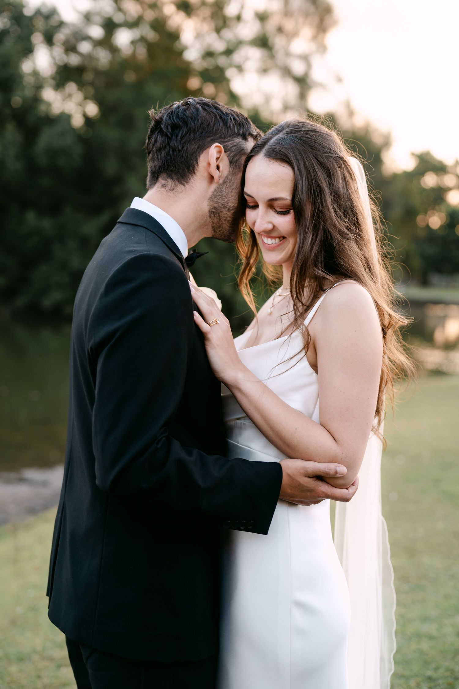 A groom in a black tuxedo tenderly embraces his smiling bride in a white gown outdoors at golden hour.